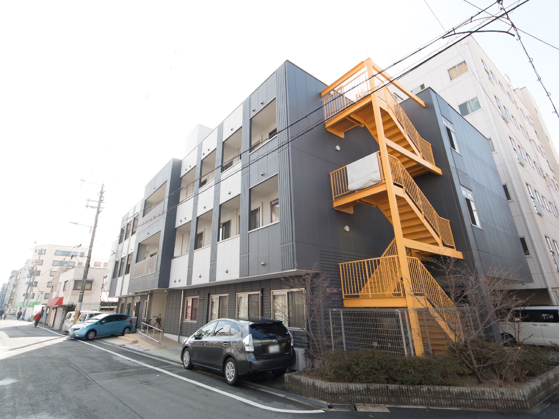 黄色にペイントされた非常階段が設置されている建物である。施設前と横には、駐車スペースが設けられている。