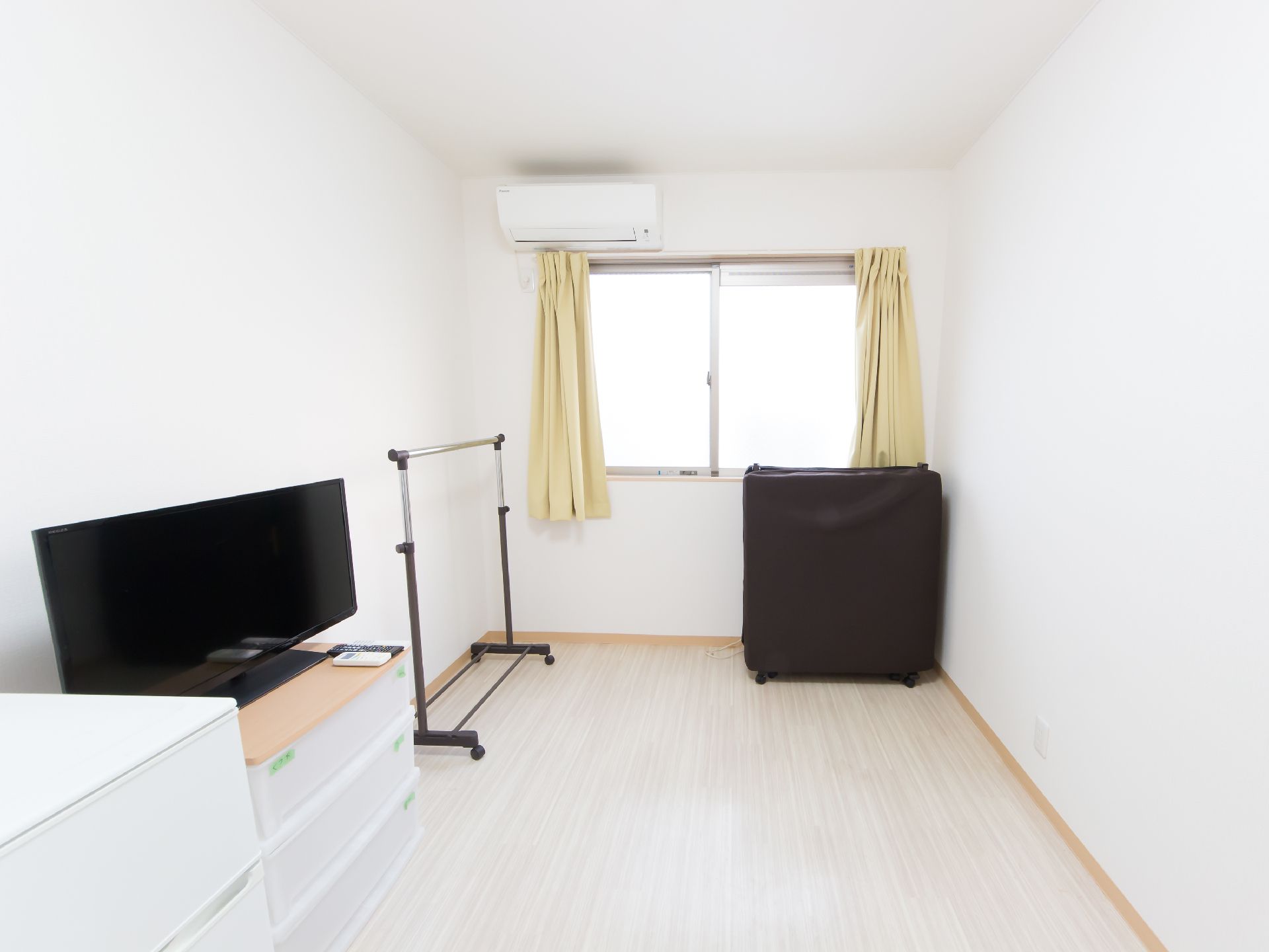 白を基調とした清潔感のある部屋で、エアコンが設置されている。窓にはカーテンが取りつけられており、折り畳み式のベッドが置かれている。