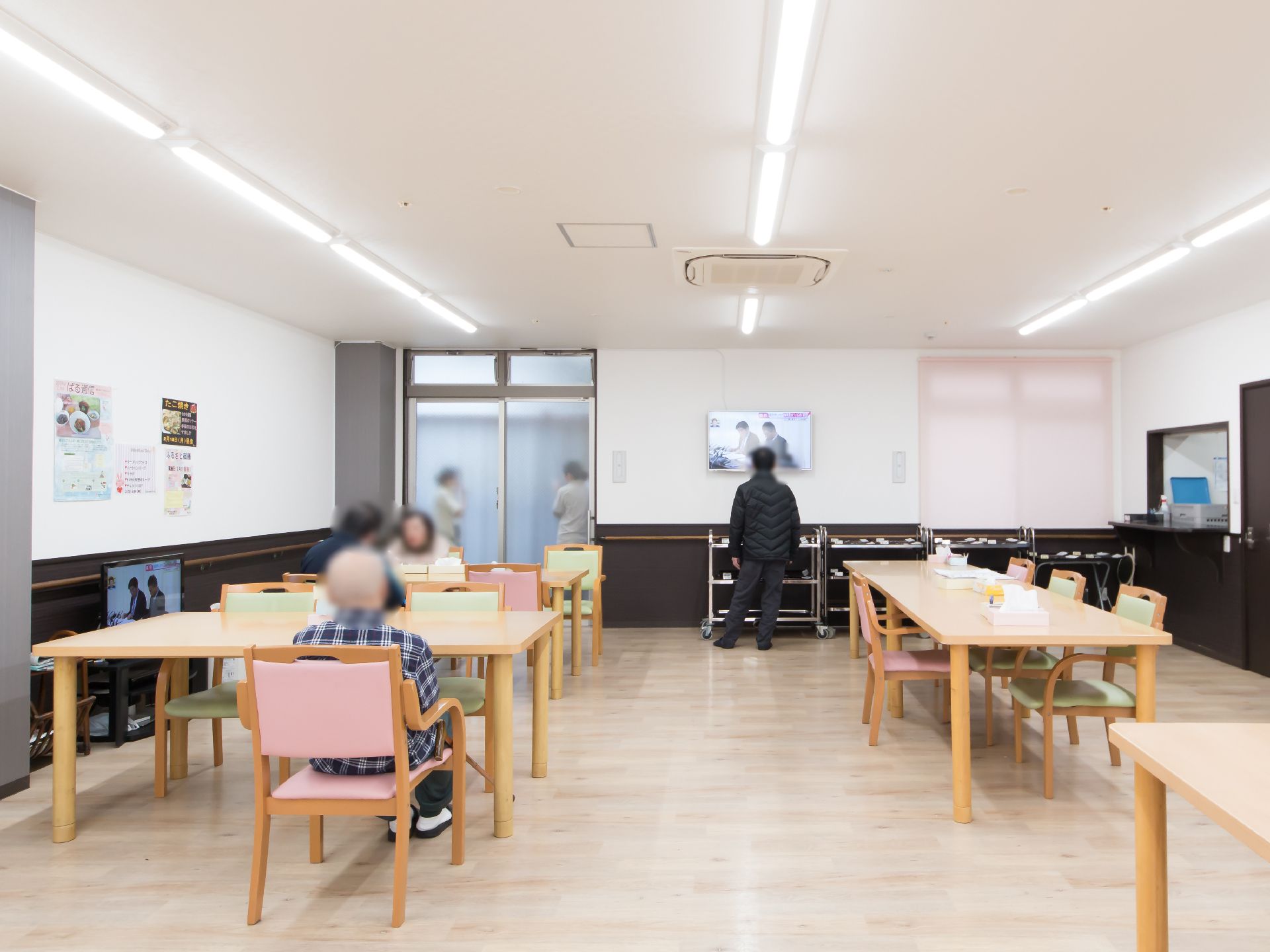 テレビが２台設置されているリビング兼食堂で、厨房が併設されている。天井には長い照明器具が３列並んでいる。