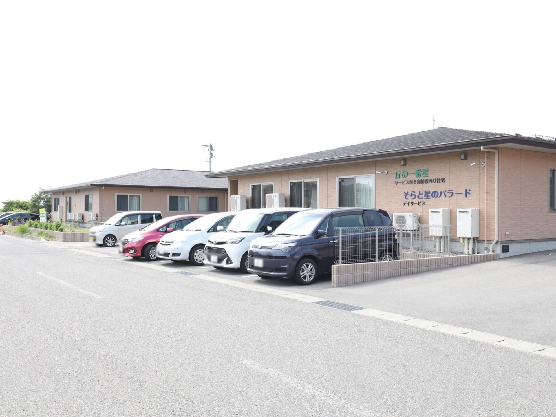 建物の前は駐車場となっていて、車が５台止まっている。外壁には施設名と併設するデイサービスの名前が掲げられている。