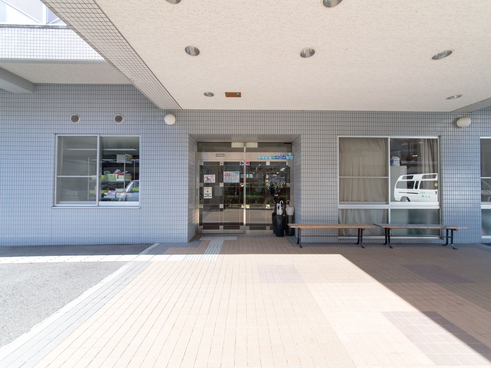 施設入口を正面から見た様子。自動ドアの前に傘立てが置かれている。また、ベンチが2つ並んでいる。玄関前にはタイルが敷かれている。