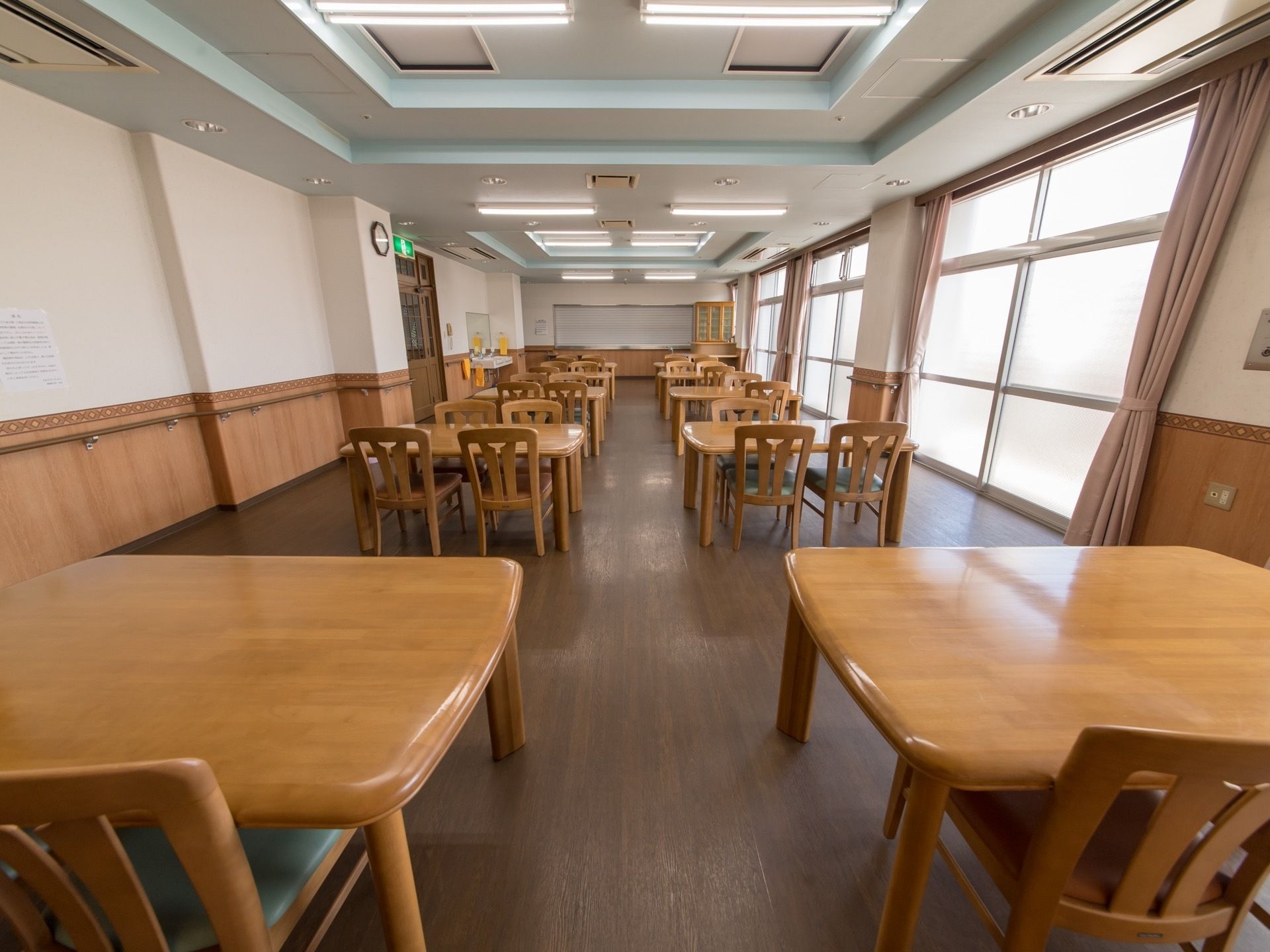 奥行きがある部屋の中に、揃いのテーブルセットが10個ほど置かれている。右側に窓が並んでいて、ピンク色のカーテンが付いている。