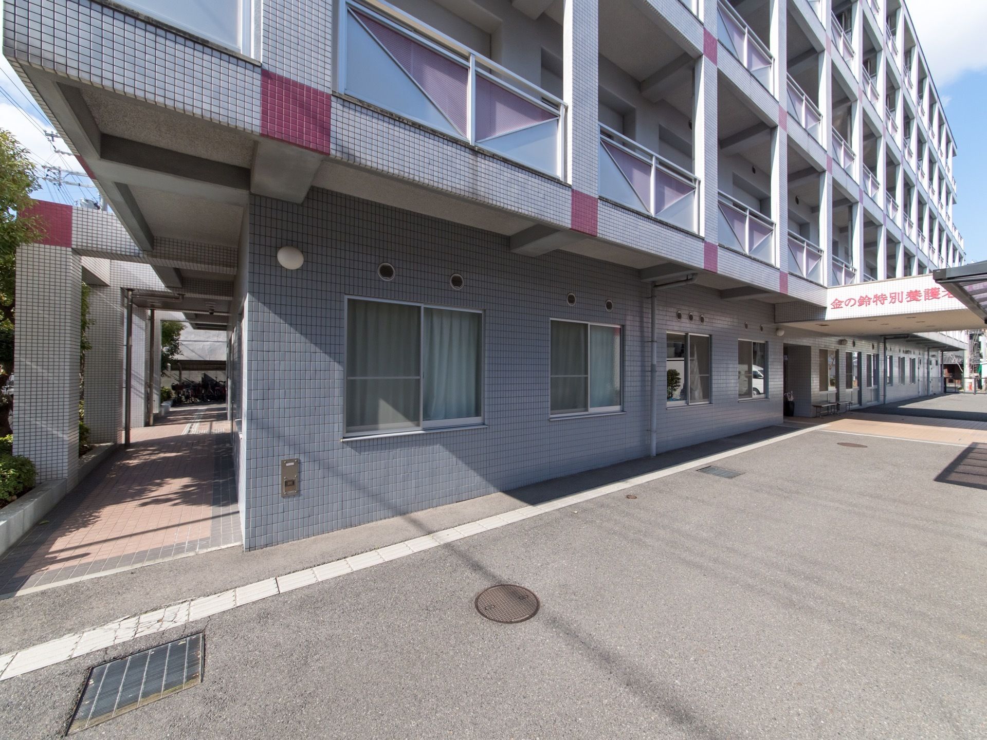 施設の建物がある。施設の前はコンクリートが敷かれ、点字ブロックが設置されている。建物の左側には、タイル敷きの通路がある。
