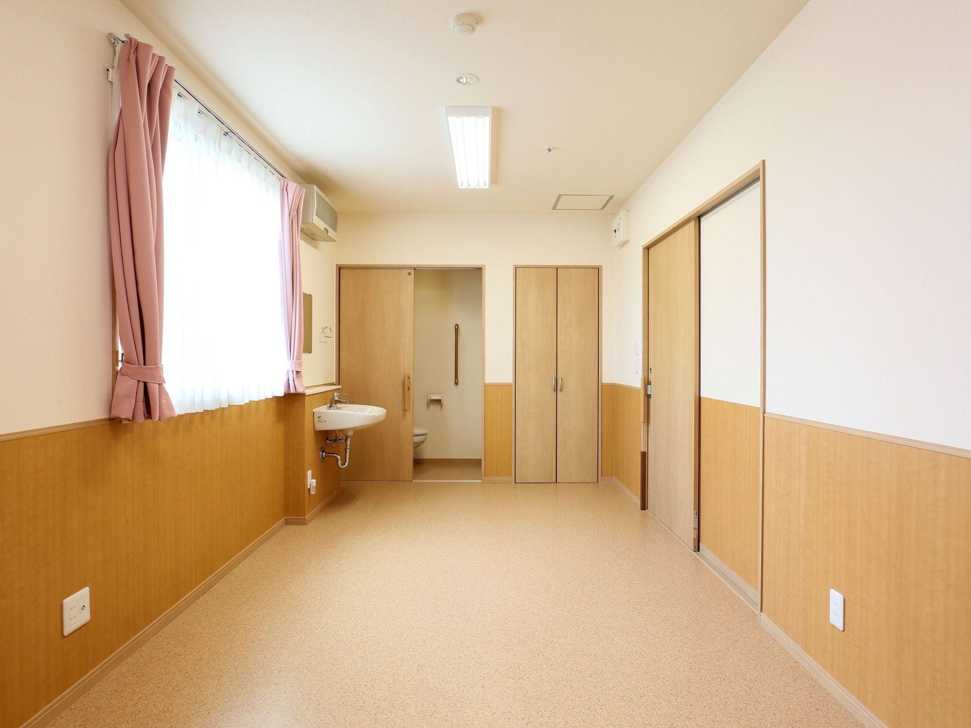 天井にライトと火災報知器が取り付けられた個室で、左側に窓がある。窓にはレースとピンクのカーテンが掛けられている。