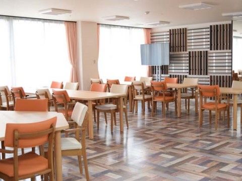 ダイニングテーブルや椅子が設置され、多くの入居者様がご利用いただける。木製の格子状になった壁があり、テレビが置いてある。