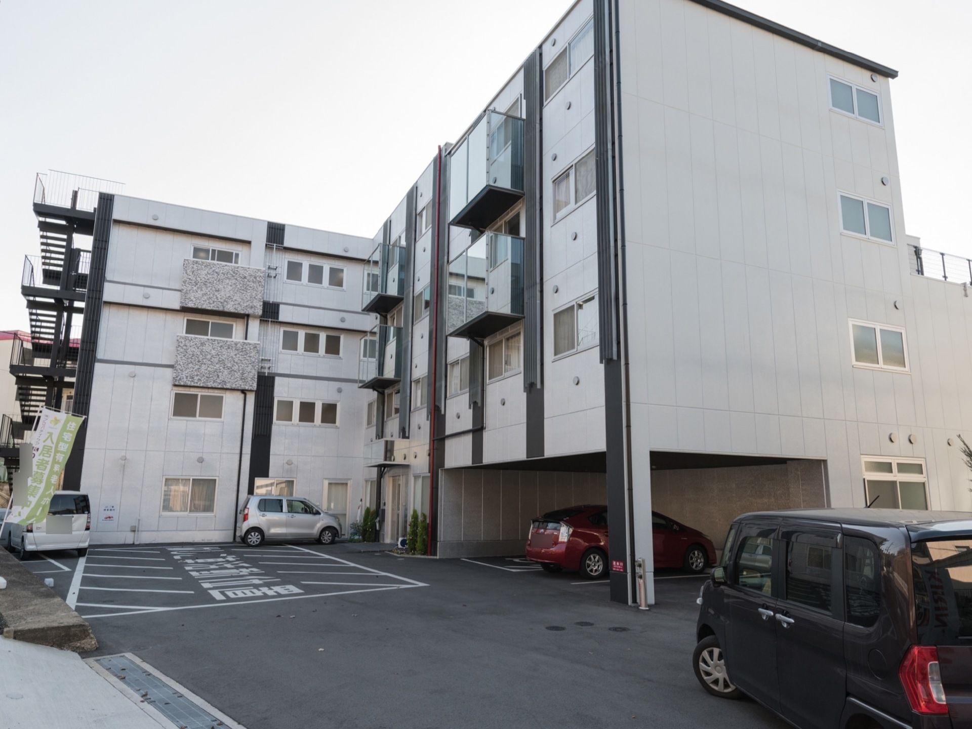 外壁が白色と縦に伸びる黒色になっている施設外観である。道路からアクセスしやすい駐車場の出入口になっている。