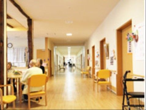 長く幅広い廊下がまっすぐ続いている。手前の一部に幅が広くなった所があり、入居者様がテーブルを囲んでお話しておられる。居室扉横に椅子がある。