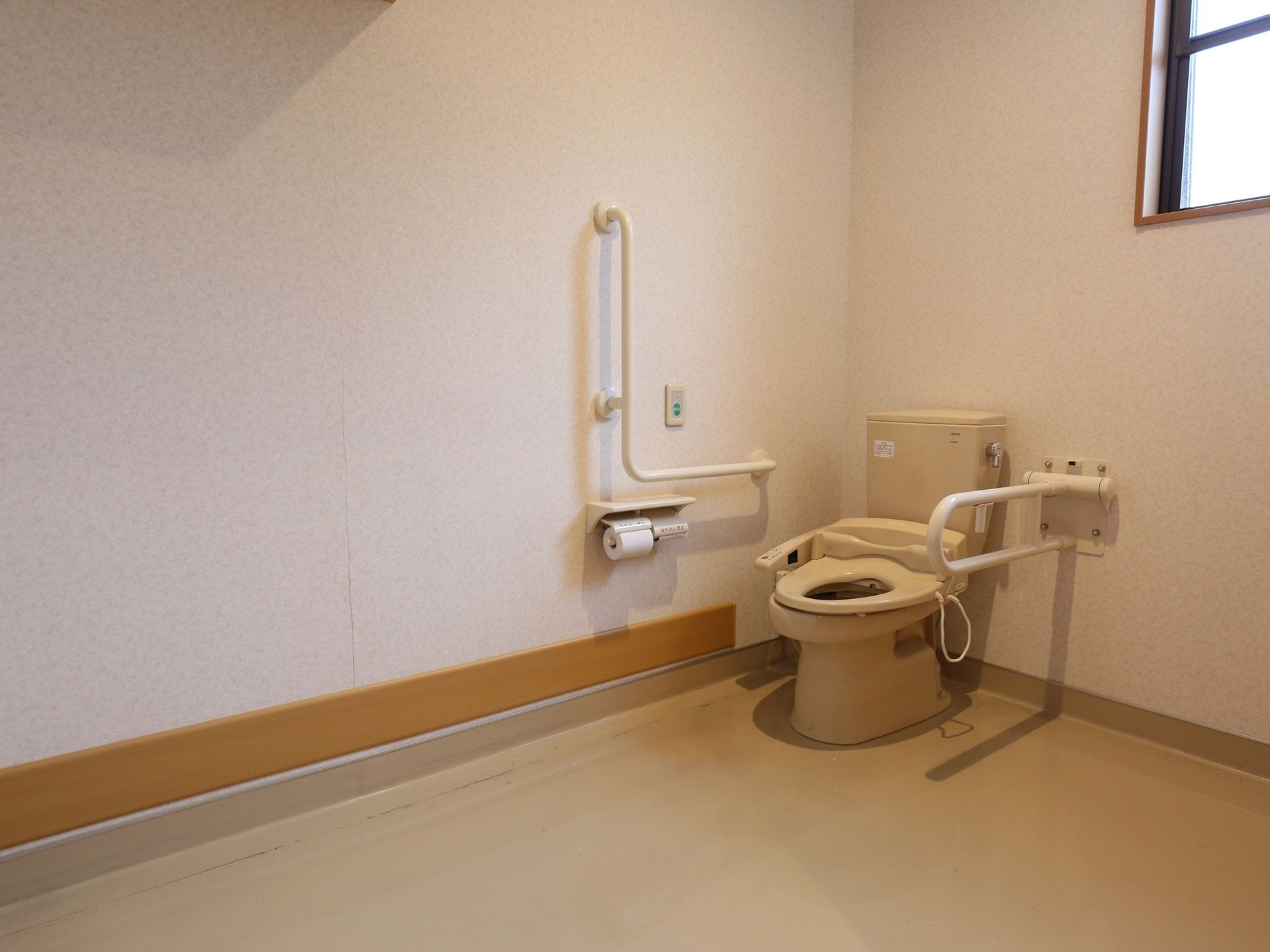 トイレは広いスペースに設置されている。壁には大きなＬ字型手すりが取り付けられている。その近くに呼び出しボタンがついている。