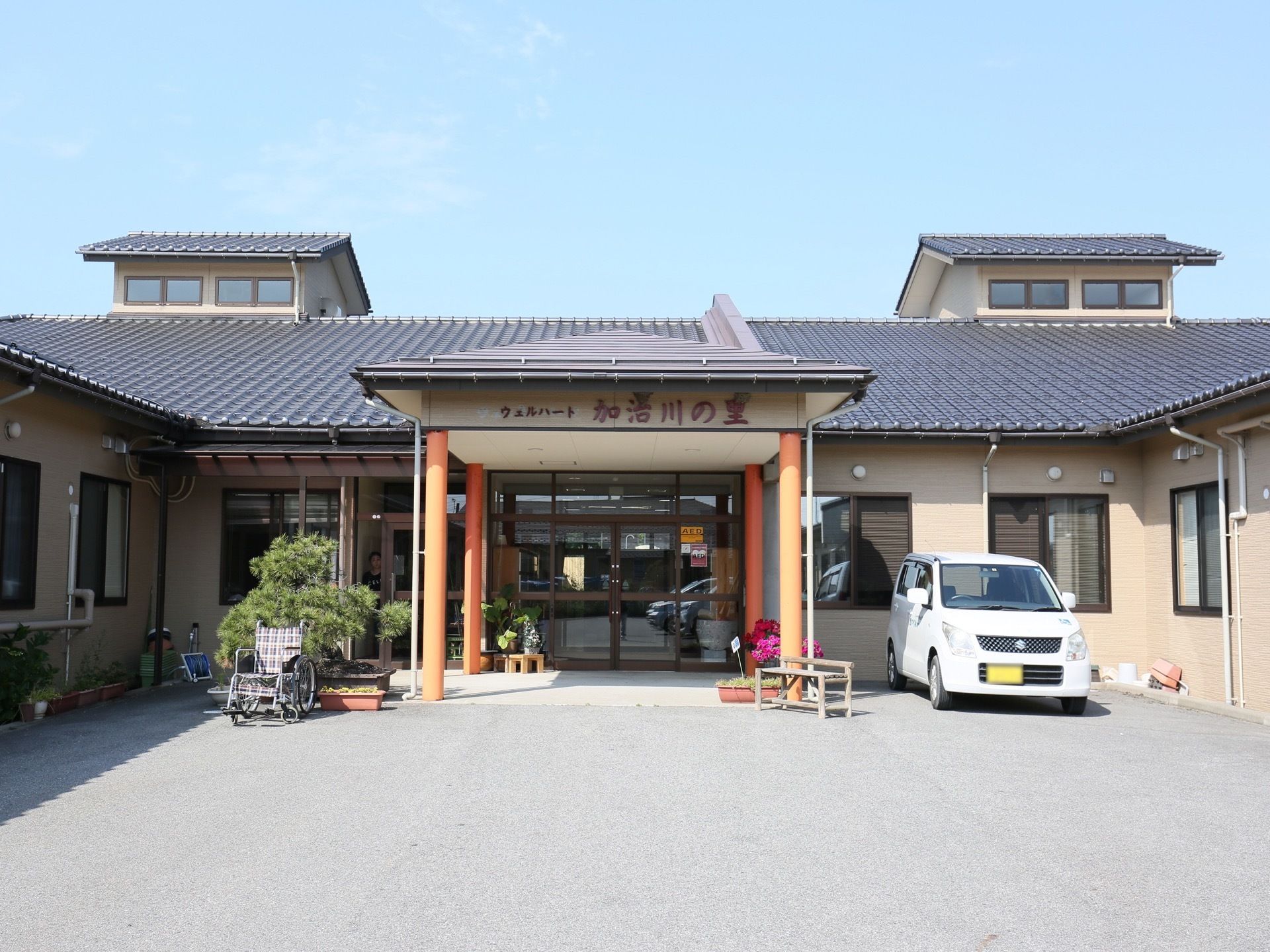 玄関には、オレンジ色の柱で支えられた大きなひさしがある。その右側に軽自動車が停まっている。左側には車椅子が置かれている。