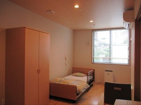 大きな窓から採光があり、明るい居室である。ベッドには転倒防止の柵がついている。洋服ダンスや空気清浄機が置かれていてくつろげる空間だ。