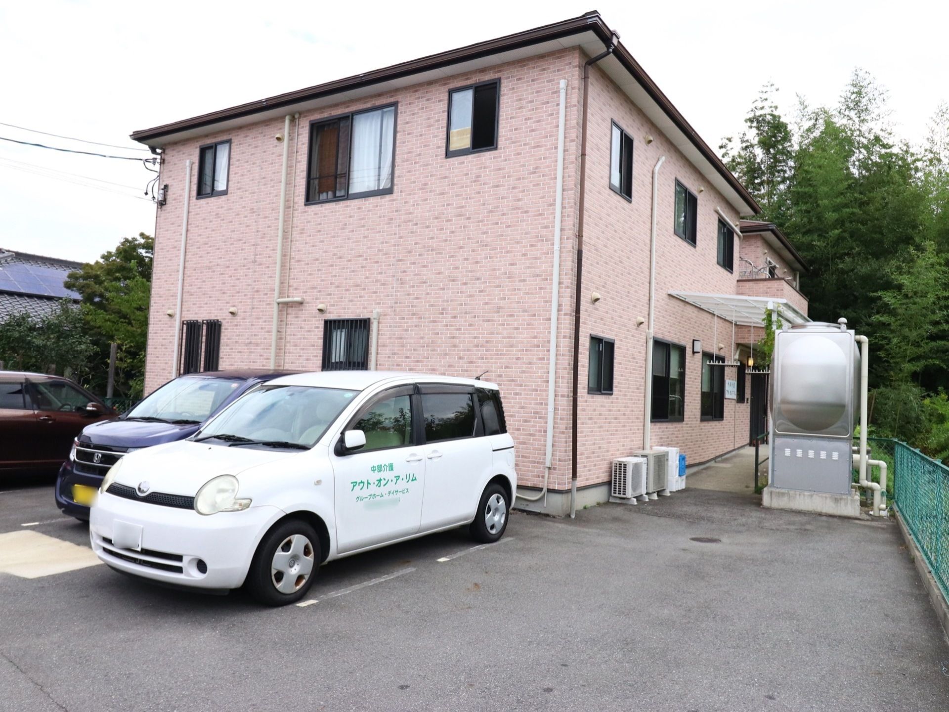 優しい茶系の外観の２階建ての建物である。建物傍には、車を停めるスペースがある。建物横にはスロープを設け横側に手すりを取り付けている。