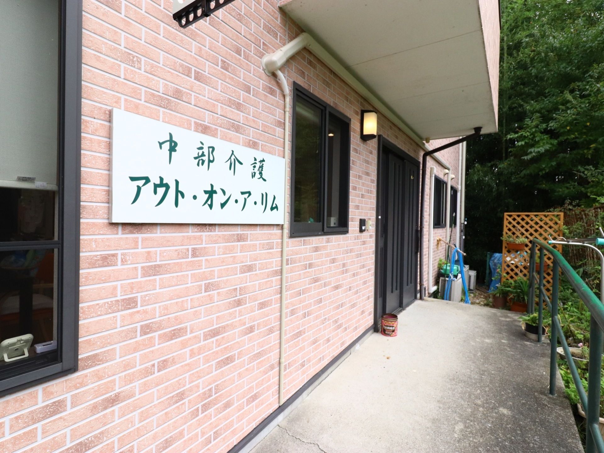 茶色のレンガ調の外壁に黒色格子の玄関ドアである。上には屋根があり、隣には、黒枠の窓がある。傍に白色の版に緑の文字の施設名の看板がある。
