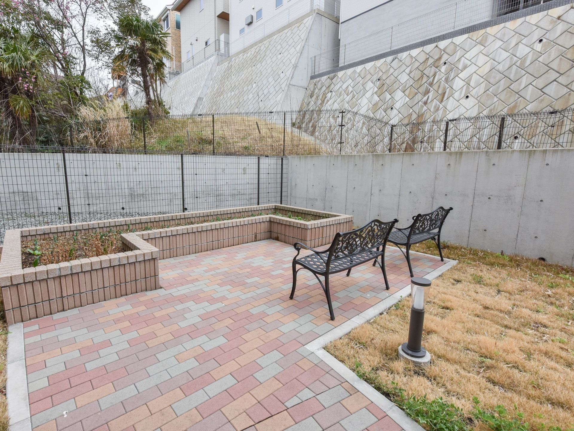 レンガ調の遊歩道の先には、大きな花壇とベンチが２脚ある。周りは芝生に囲まれていて、ライトが設置されている。