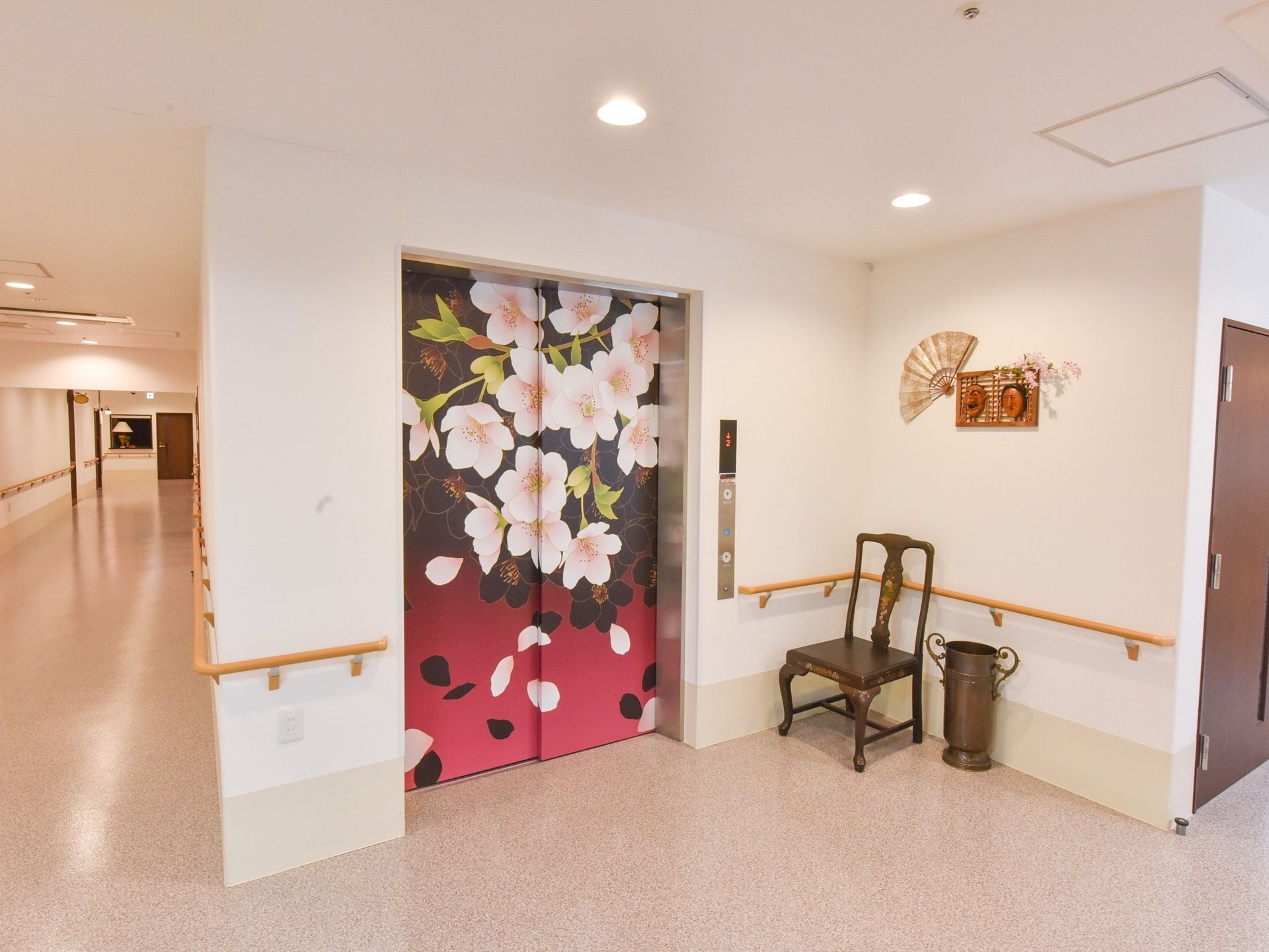 桜の花が描かれたエレベーターの前には、椅子が置かれ、壁には手すりが設置されている。壁にも和を感じられる装飾がされている。