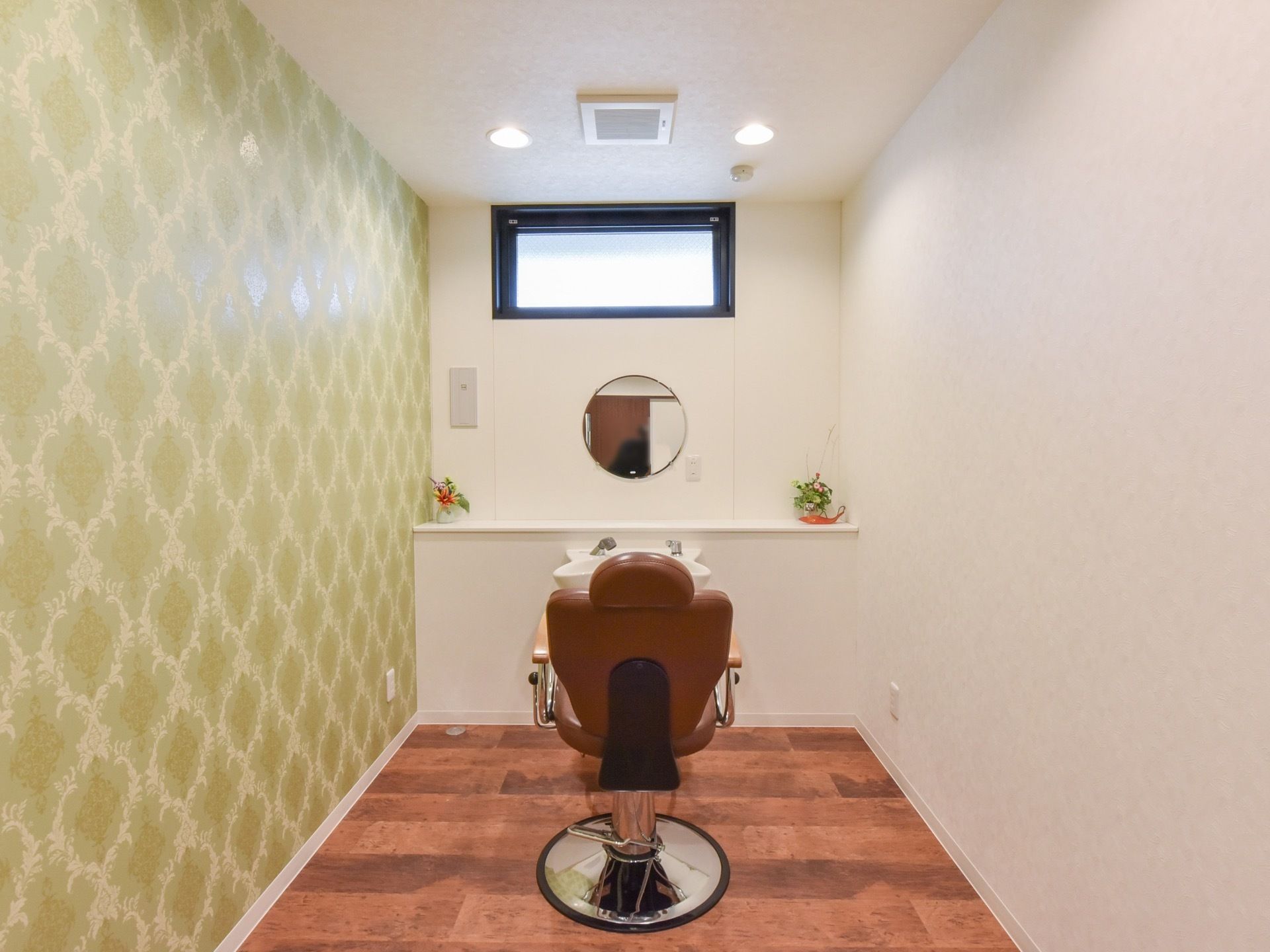 お洒落な個室のヘアサロンである。ゆったりと座れる椅子の前には、洗面台と丸い鏡があり、花が飾ってある。