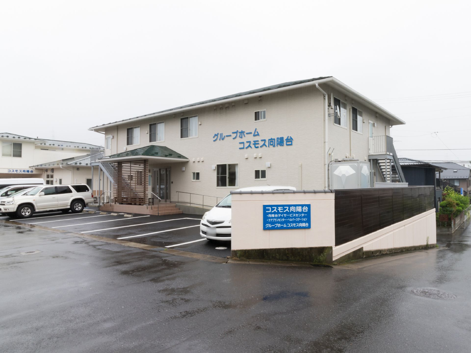 閑静な住宅街にある施設の様子。建物の壁には施設名が記されている。充分な駐車スペースが設けられている。玄関はバリアフリー。