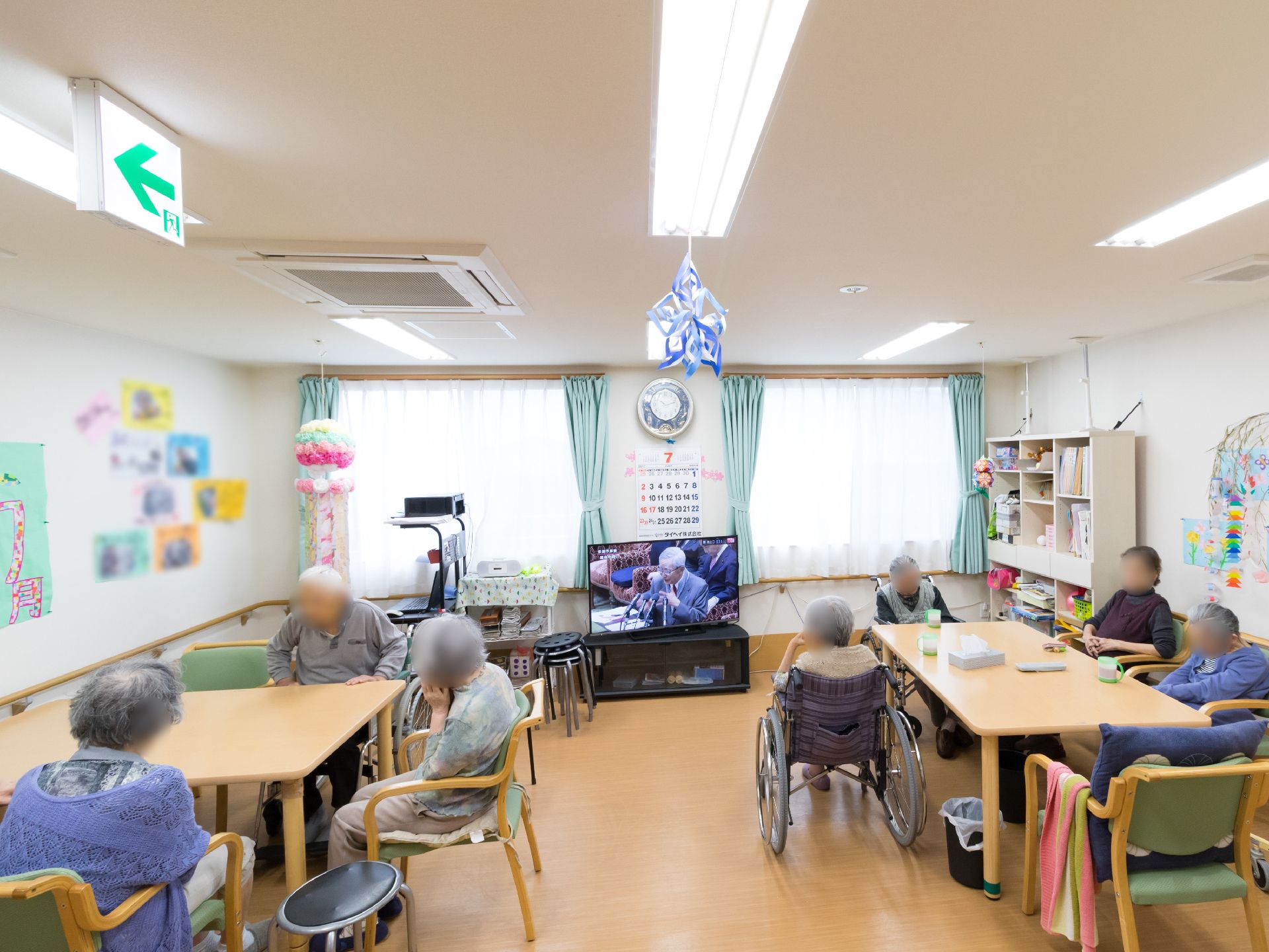リビングにはテレビがあり、居住者たちはテレビ鑑賞を楽しんだり、会話を楽しんだりしている。テーブルや椅子があり、車椅子の居住者もいる。