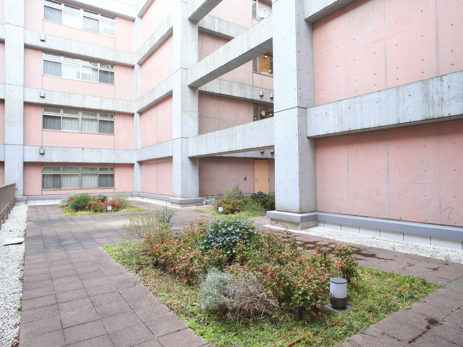 施設の建物に囲まれた中庭。植栽スペースが数か所あり、その周りを茶色の四角いブロックで、その周りを白い砂利で囲まれている。