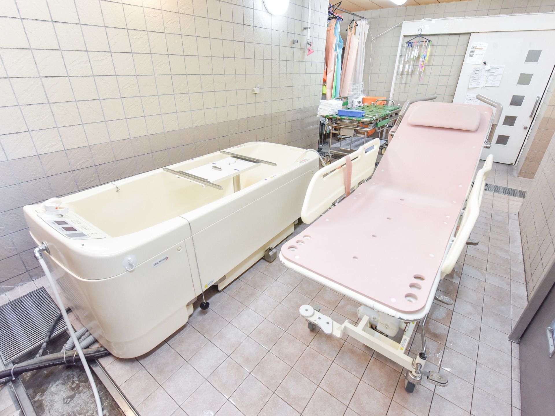 奥に引き戸の白い扉がある。機械浴用の浴槽が壁際に、隣に浴槽に浸かれるベッドがある。浴槽は白色である。ベッドはピンク色で両横に手すりがある。