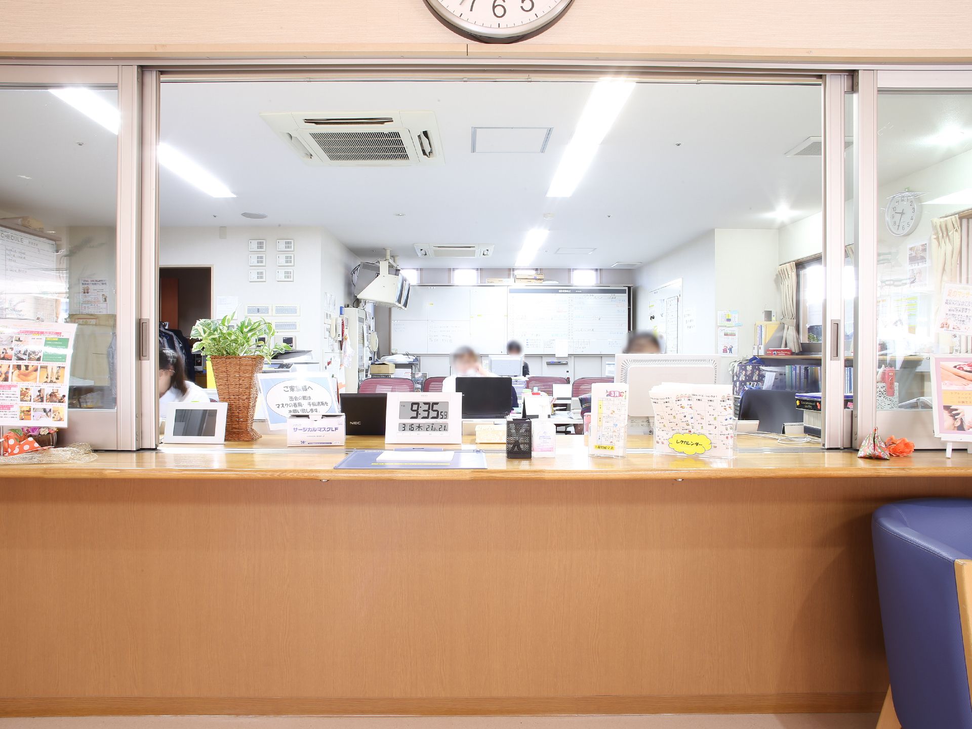 幅広い受付カウンターには、案内文書やデジタルの時計が置かれている。カウンターの奥はスタッフルームで、机で事務仕事をしている様子が見える。