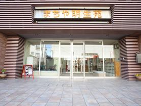 エントランスのポーチの壁に黄色い字で施設名が大きく表示されている。自動ドアの両側には花が飾られている。