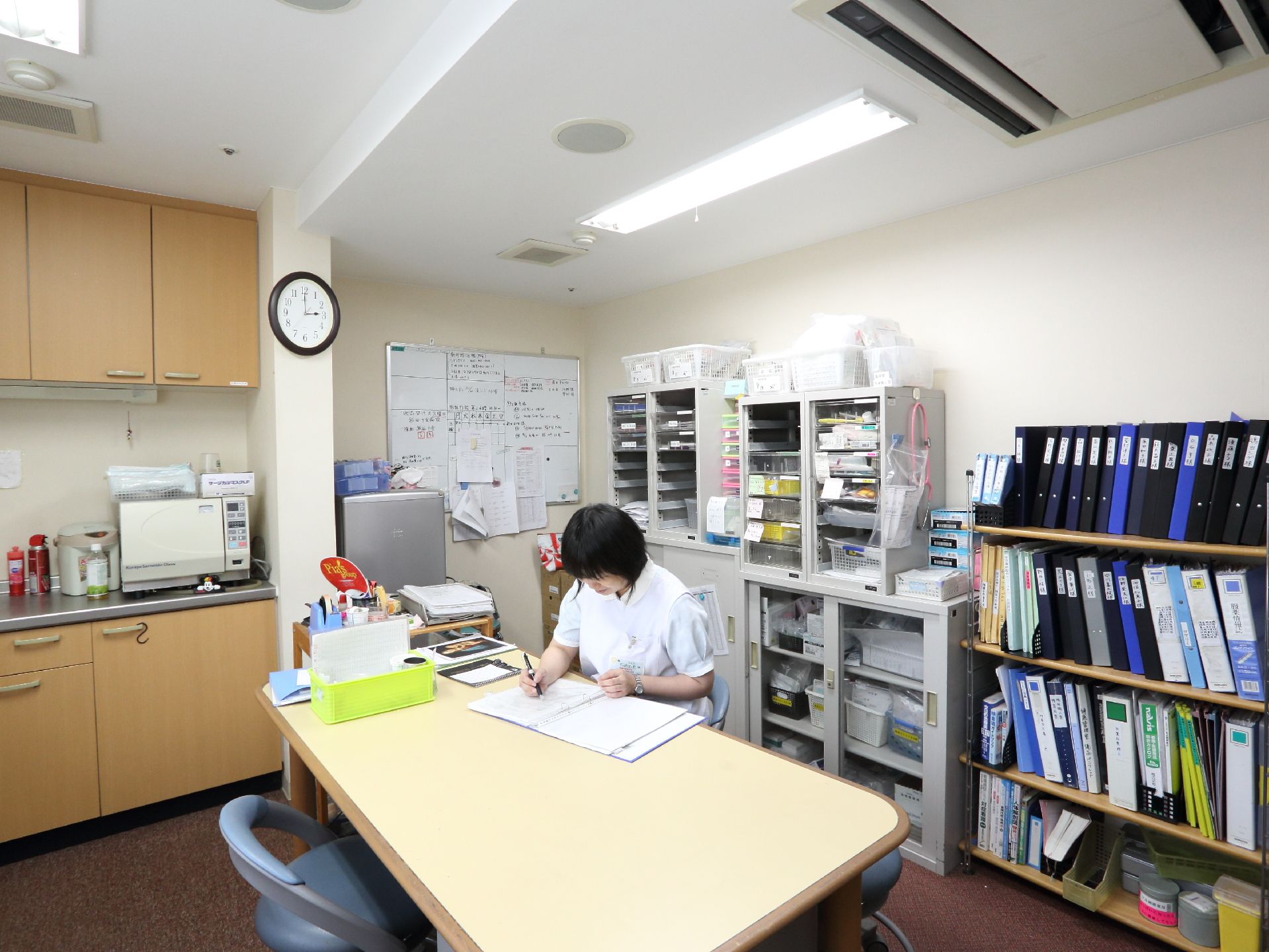 スタッフが打ち合わせや作業を行う部屋。中央に机と椅子が置かれ、壁際には本棚や書類棚が整然と並べられている。