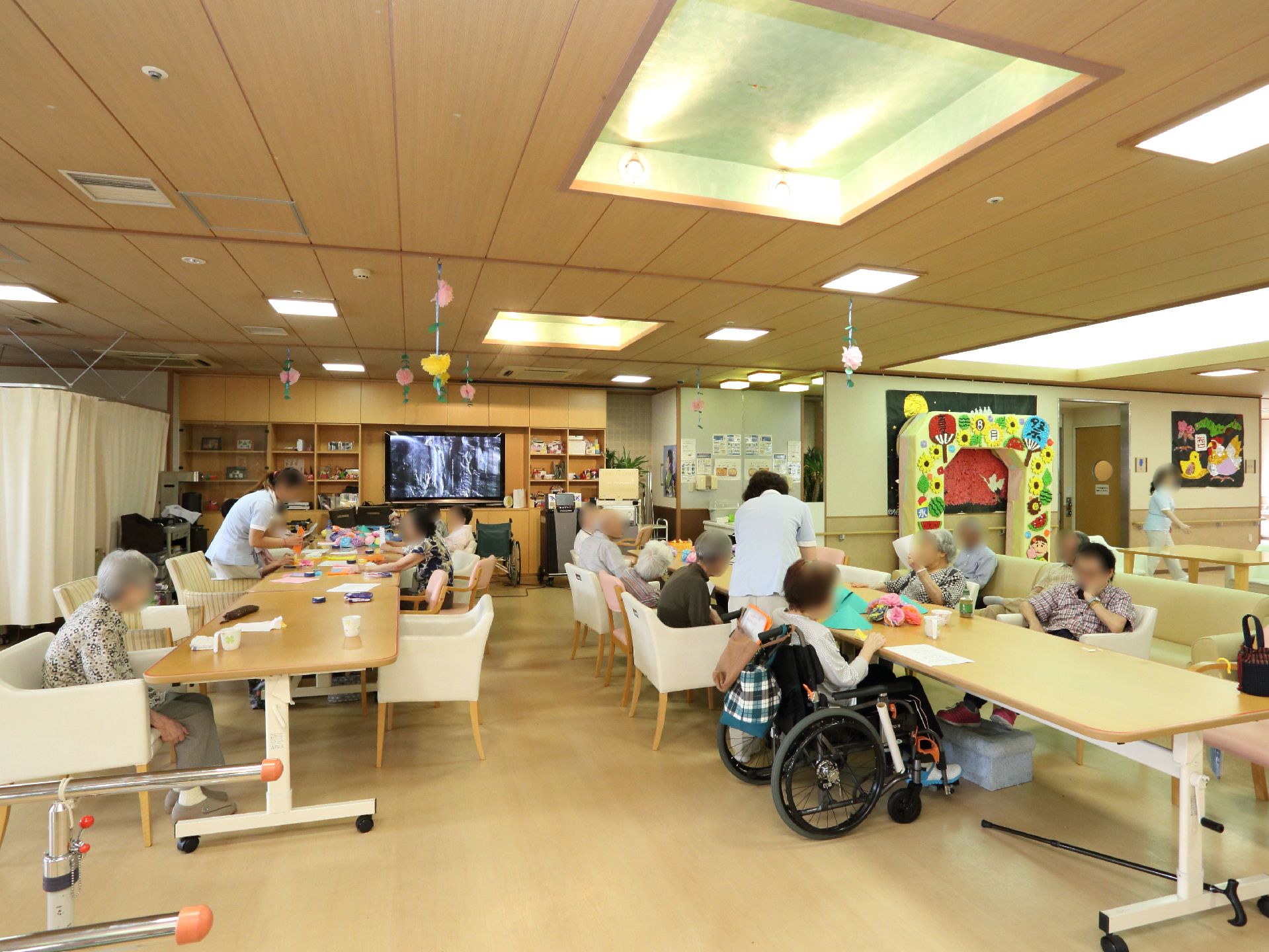 開放的なリビングには、あちこちにご利用者様の手作りの作品が飾られている。長いテーブルと椅子が設けられている。ワイド型のテレビもある。