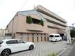 ベージュ色の壁の施設の外観。建物が車道より奥まった所に建っているので、安心できる。周りは静かな住宅街である。