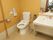 ベージュ色の壁のトイレには、タンクがついたトイレと横幅の広い洗面台が設置されている。トイレの横には、手すりも取りつけられている。