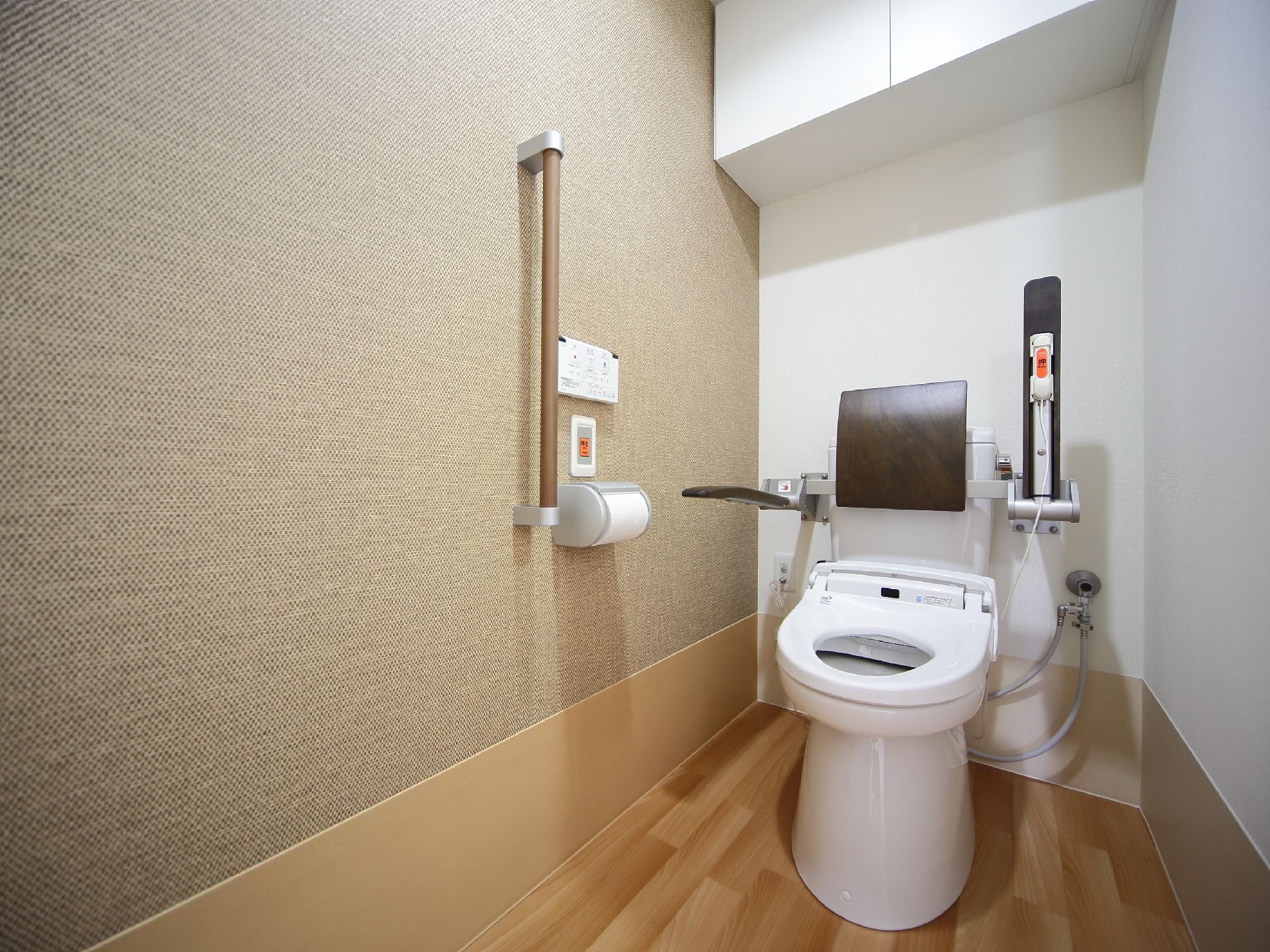 壁のパネルで操作する温水洗浄便座が設置されたトイレである。パネルの下に非常ボタンが取りつけられ、その下にトイレットペーパーがある。