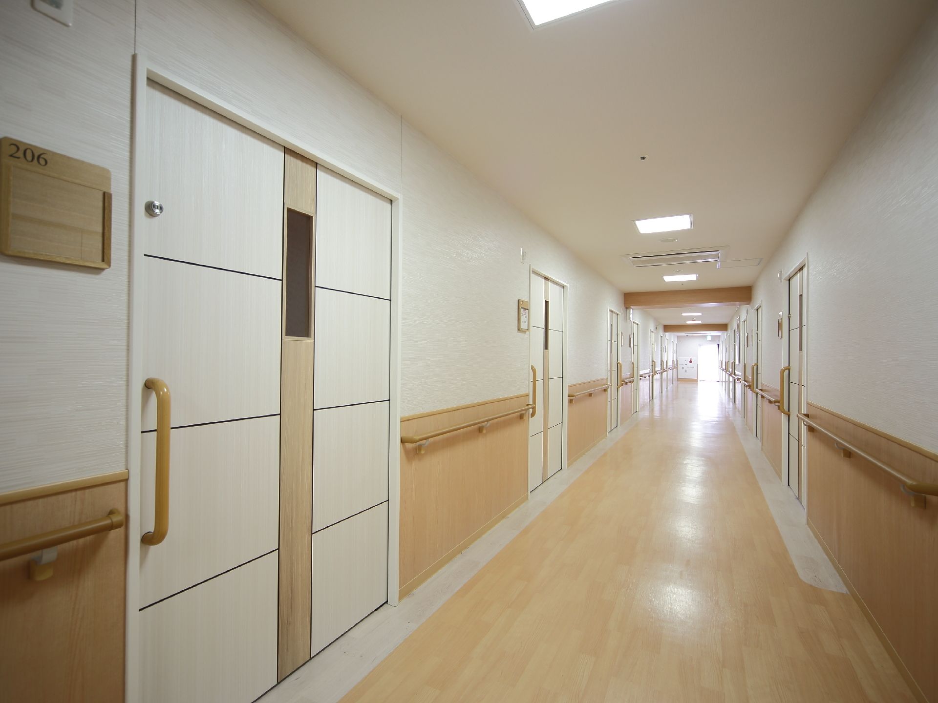 木材と白を基調とした優しい色合いの廊下である。等間隔に居室へのドアが並んでいる。壁には手すりが取りつけられている。