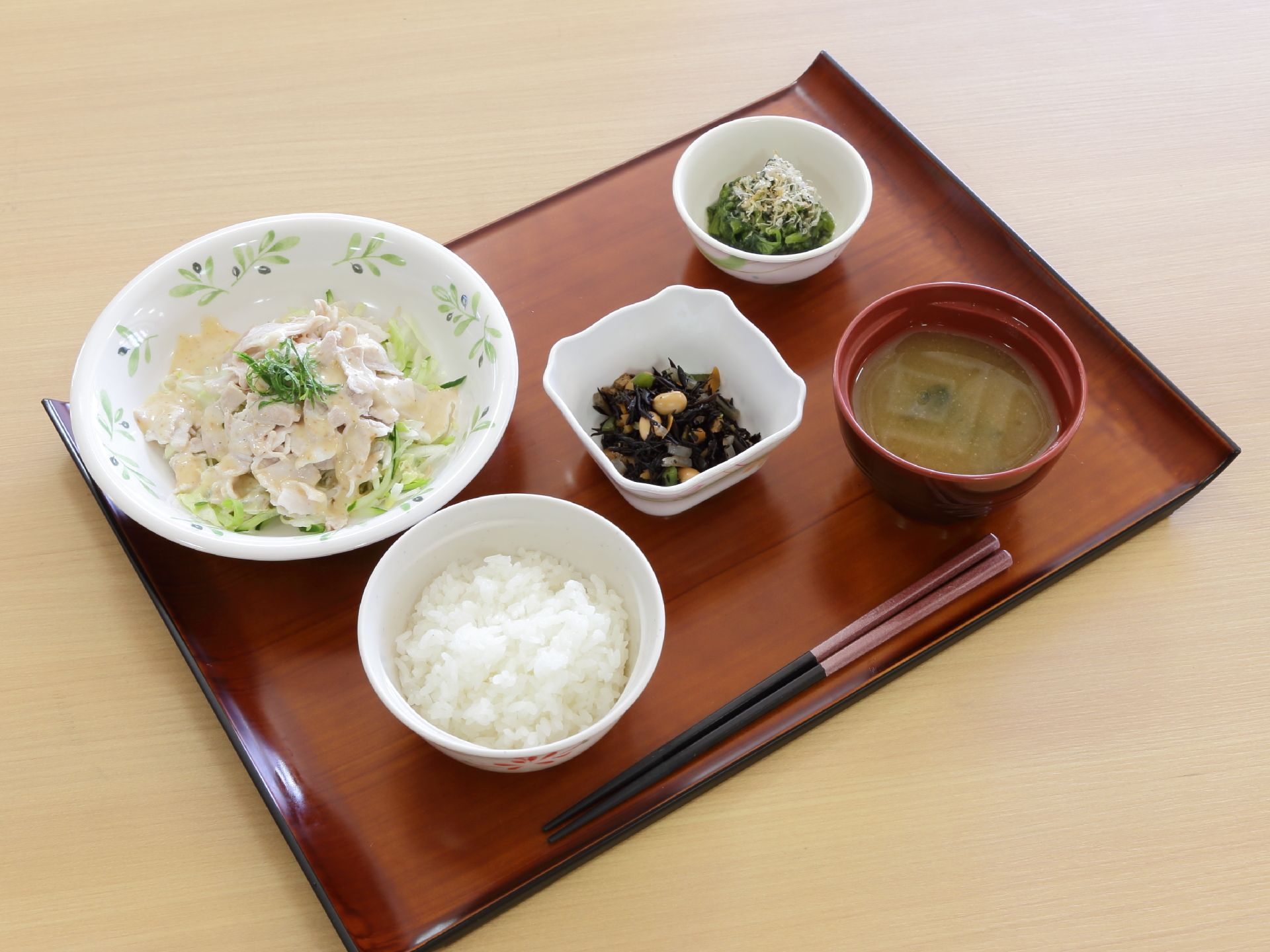 茶色のトレイに食事が乗せられている。中央にひじきの小鉢があり、サラダ、おひたし、ご飯とみそ汁がある。