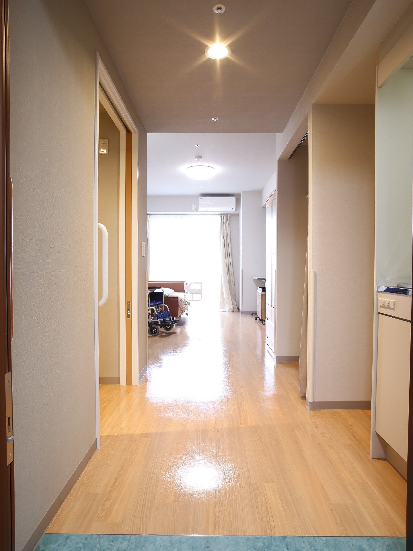フローリングの広い居室で、ミニキッチンや収納、ベッド、トイレの入り口などが見えている。ベッドの近くに車椅子がある。