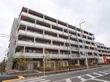 ６階建ての大きなビルのような建物が建っている。建物の前は点字ブロックが設置された広い歩道と横断歩道がある。