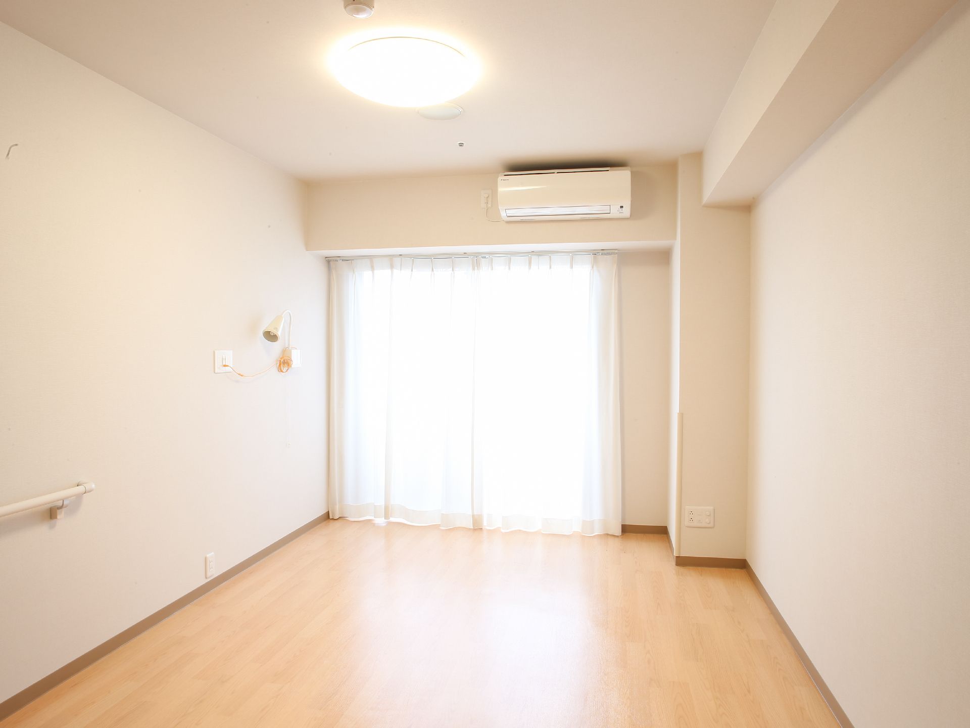 居室の奥には、大きな窓があり、カーテンがついている。窓の上には、エアコンが設置されている。また、壁には手すりがついている。