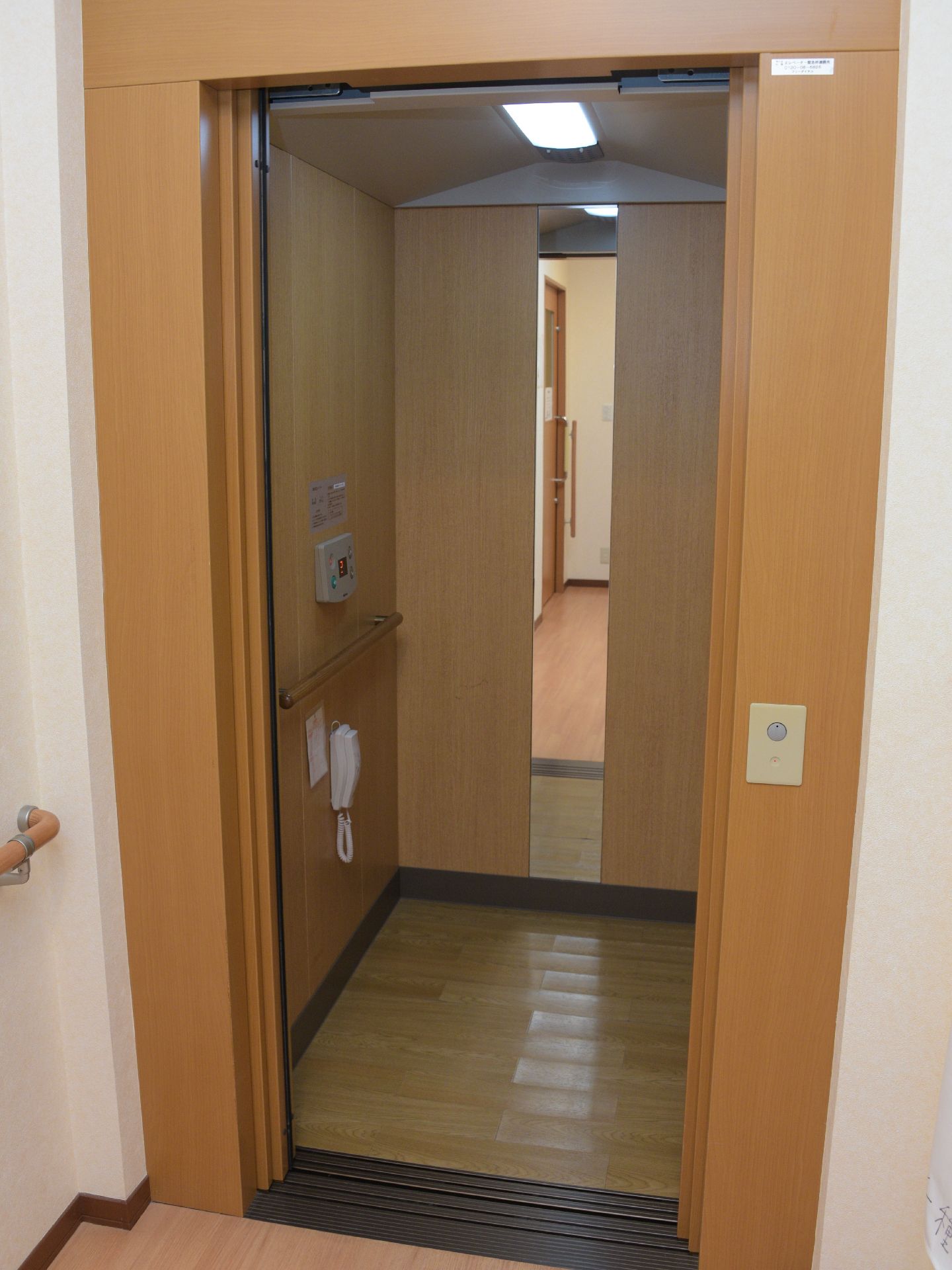 施設内のエレベータ。落ち着いた茶色の色調で、鏡がついており、車いす利用者も安全に乗り降りできるコンパクトで機能的なエレベーター。