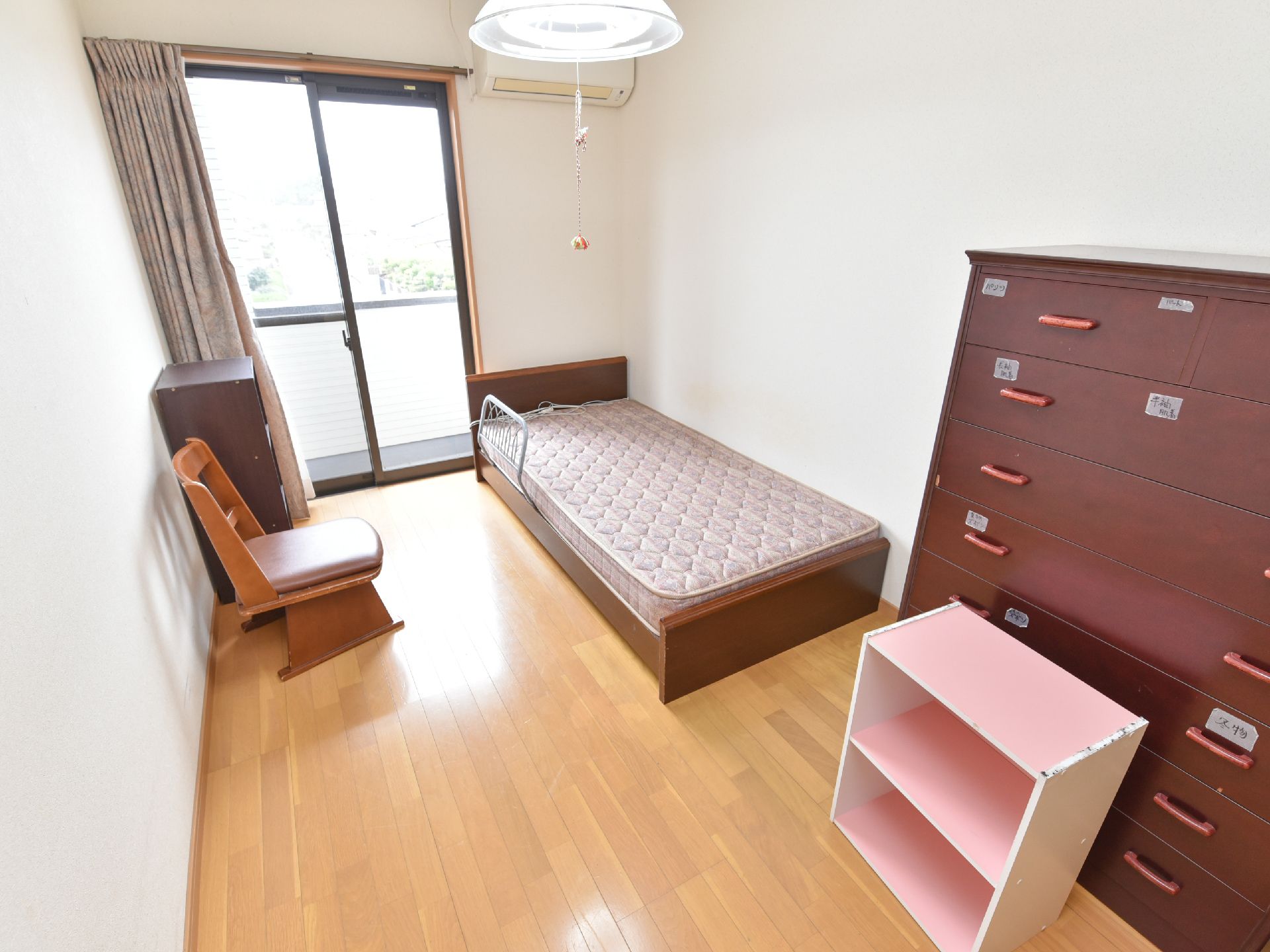 白い壁と茶色の家具で構成された居室である。タンスの引き出しには、中身がわかるようシールが貼られている。