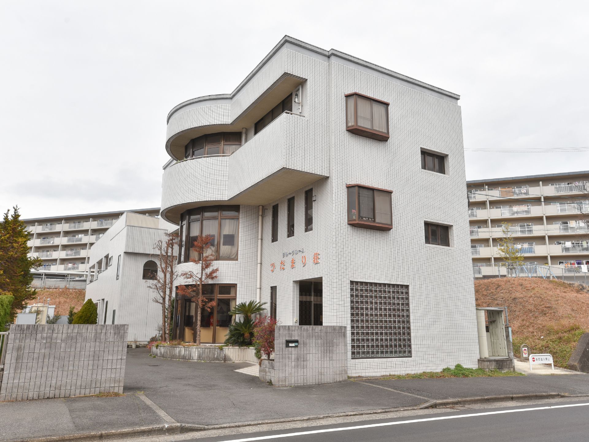 白い外壁の３階建ての建物で、玄関の上部にオレンジ色で施設名が書かれている。施設の奥にマンションが建てられている。
