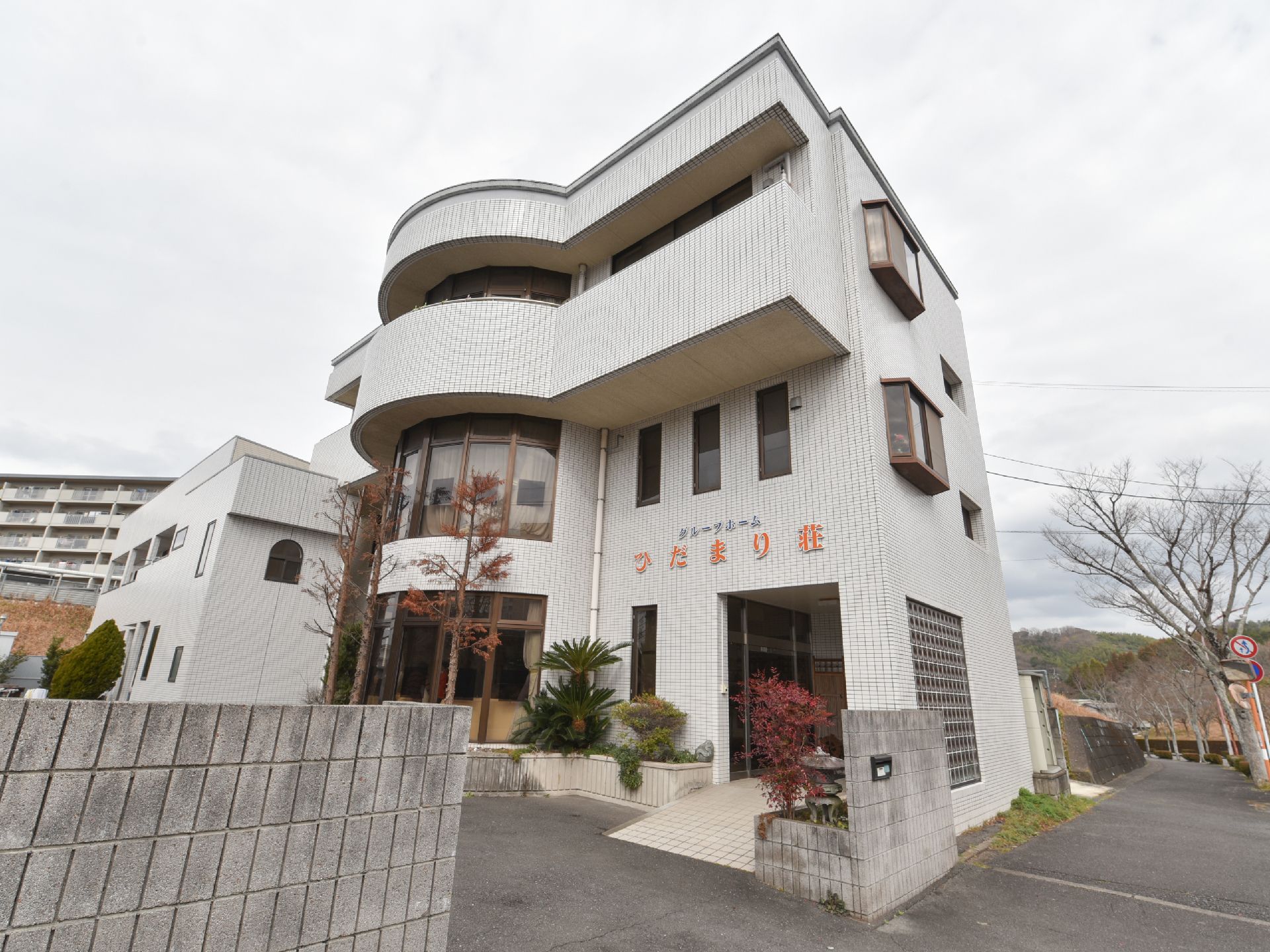 白い外壁の３階建ての建物で、バルコニーなどが設けられている。施設前は幅の広い歩道になっており、道路沿いに木々が植えられている。
