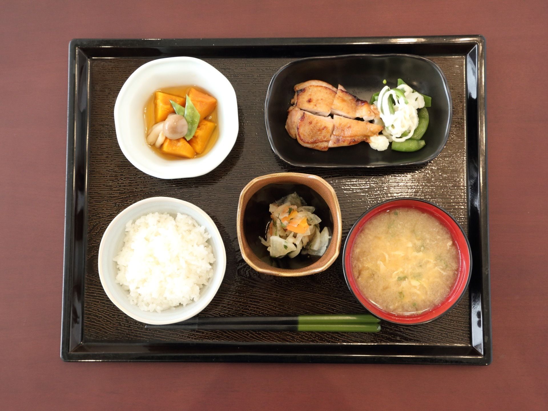 お盆の上には、メインの肉料理、カボチャの煮物、大根や人参が入った小鉢、ごはんと味噌汁がのせられている。