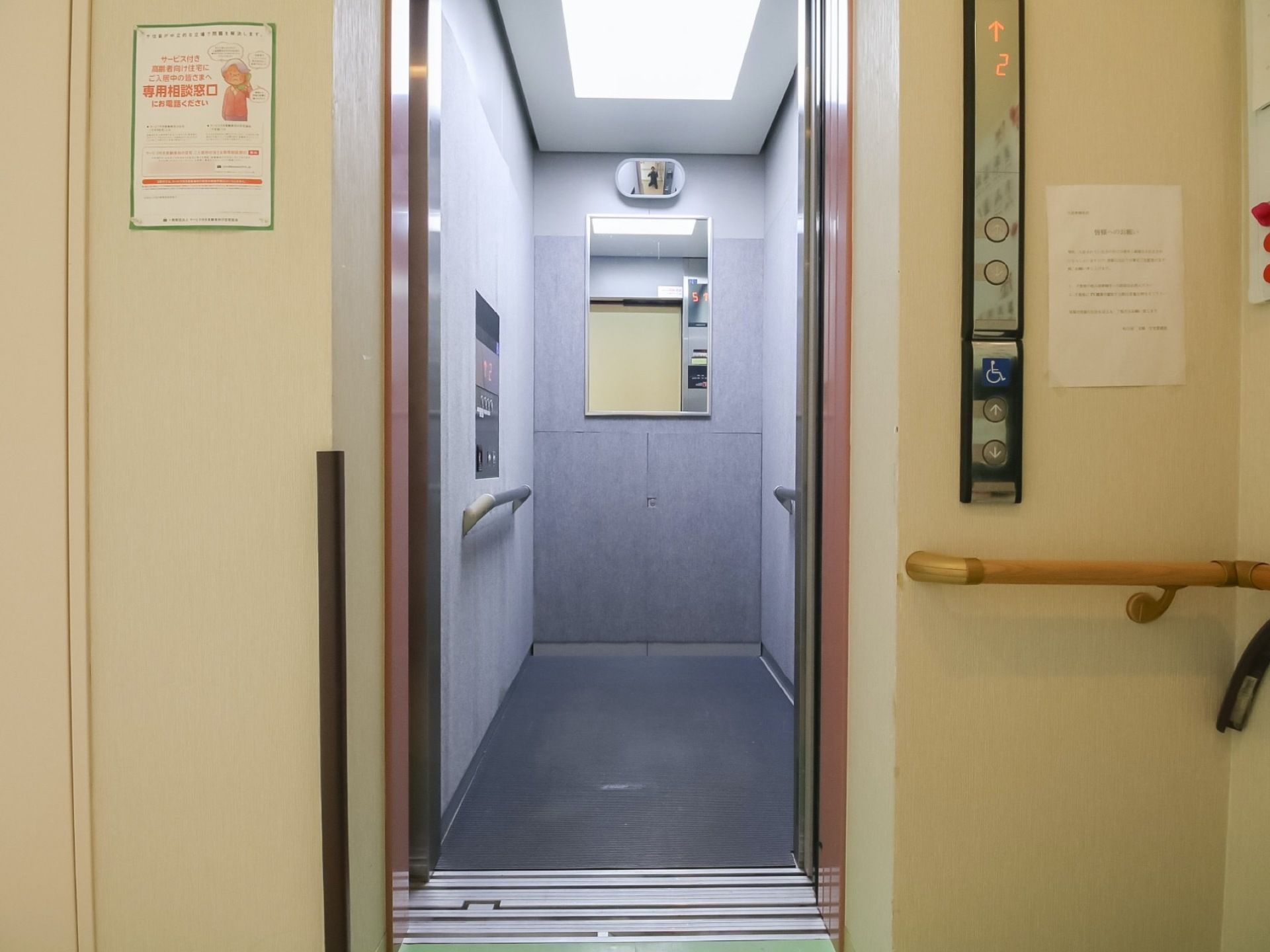 グレーの内装のエレベーターがある。正面と上部分には鏡が付いており、両側の壁には手すりが設置されている。
