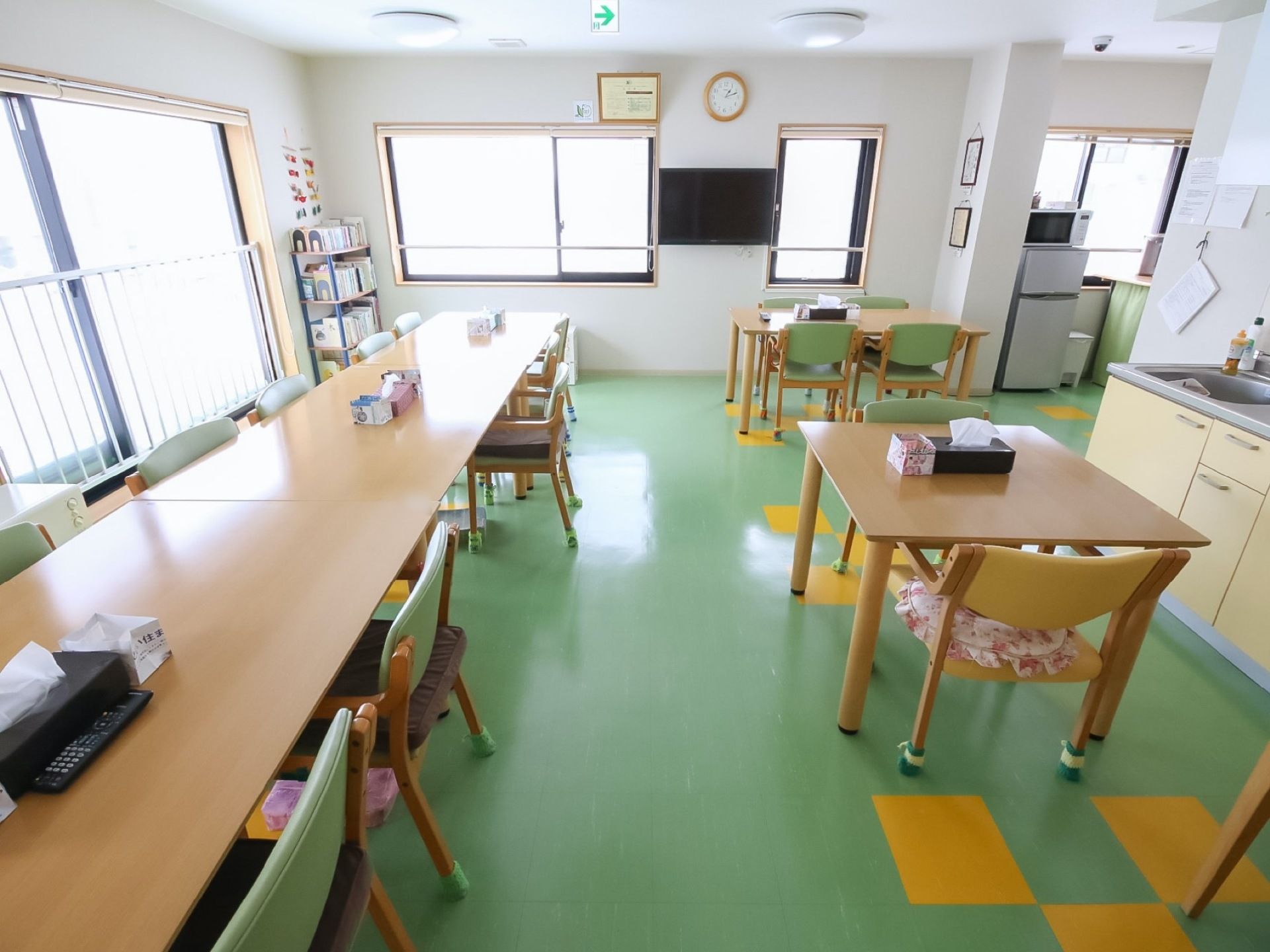 緑色の床の広い食堂がある。テーブルと椅子がたくさん並んでおり、壁には窓が付いている。キッチンがあり、本棚が置かれている。