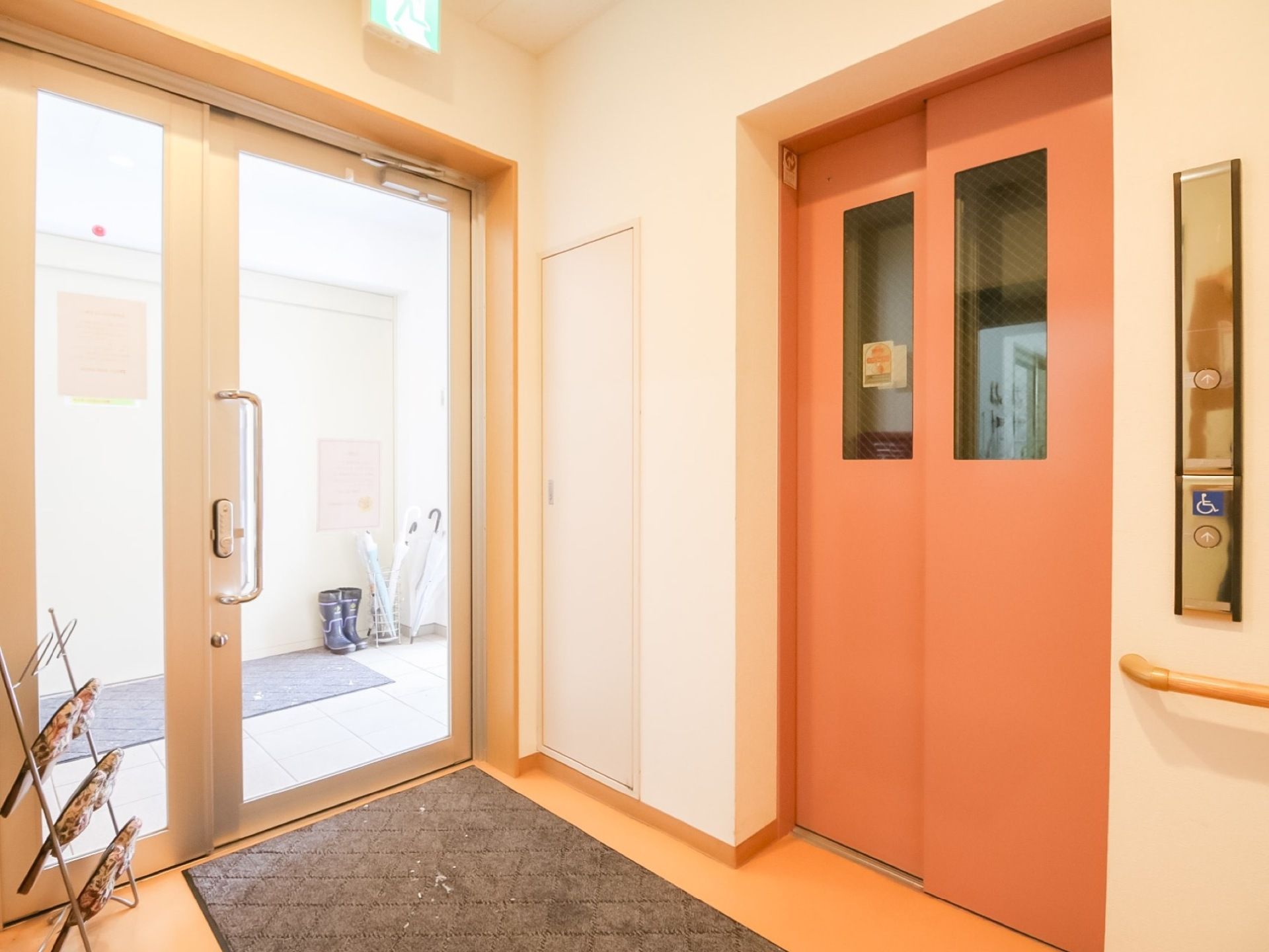 玄関を入った横にあるエレベーターホール。サーモンピンクの扉には小窓が付いている。玄関へと通じるガラス扉がある。