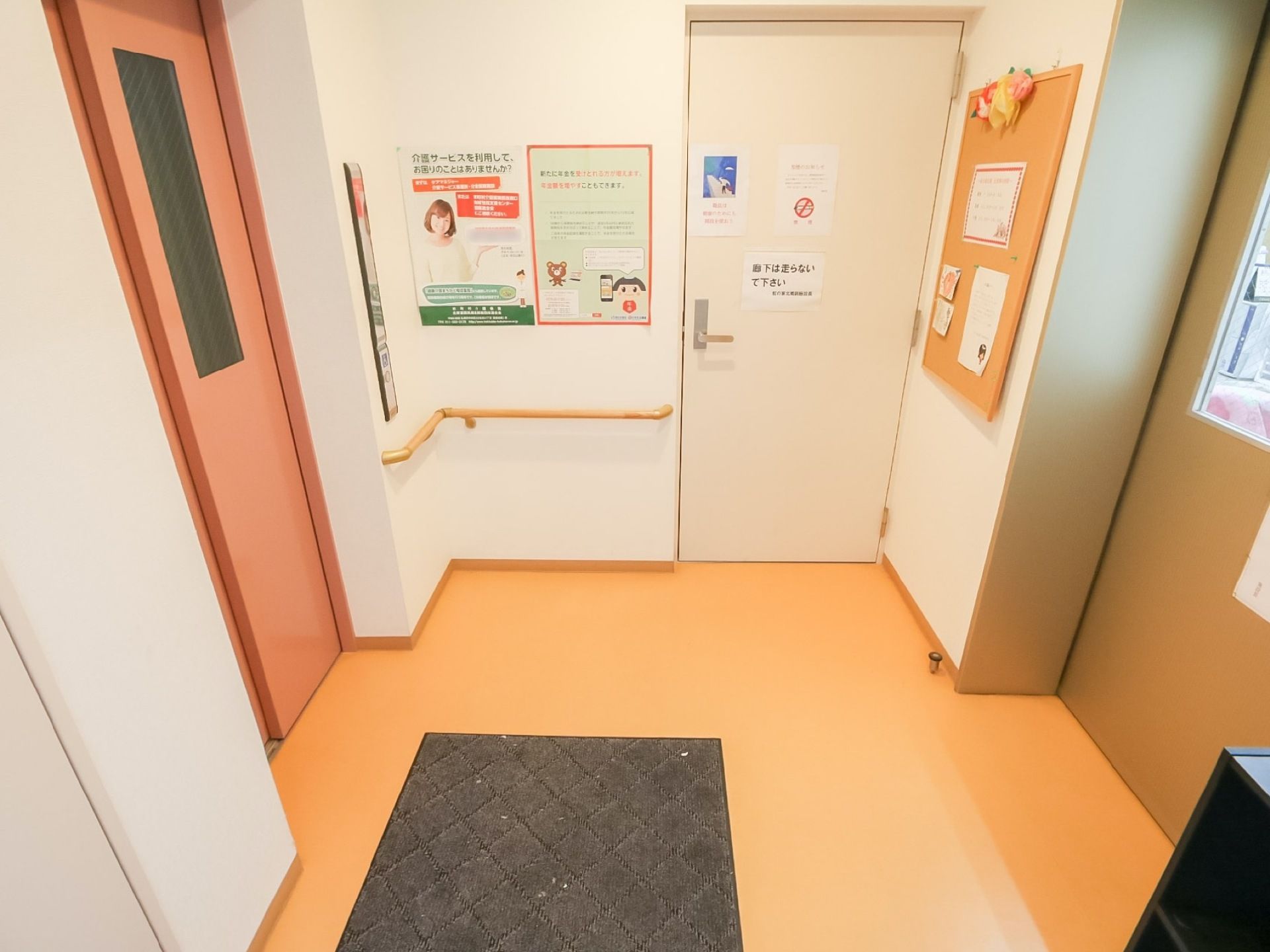 サーモンピンクのドアのエレベーターがある。地面にはマットが敷かれ、周囲の壁にはコルクボードとポスターが貼られ、手すりが設置されている。
