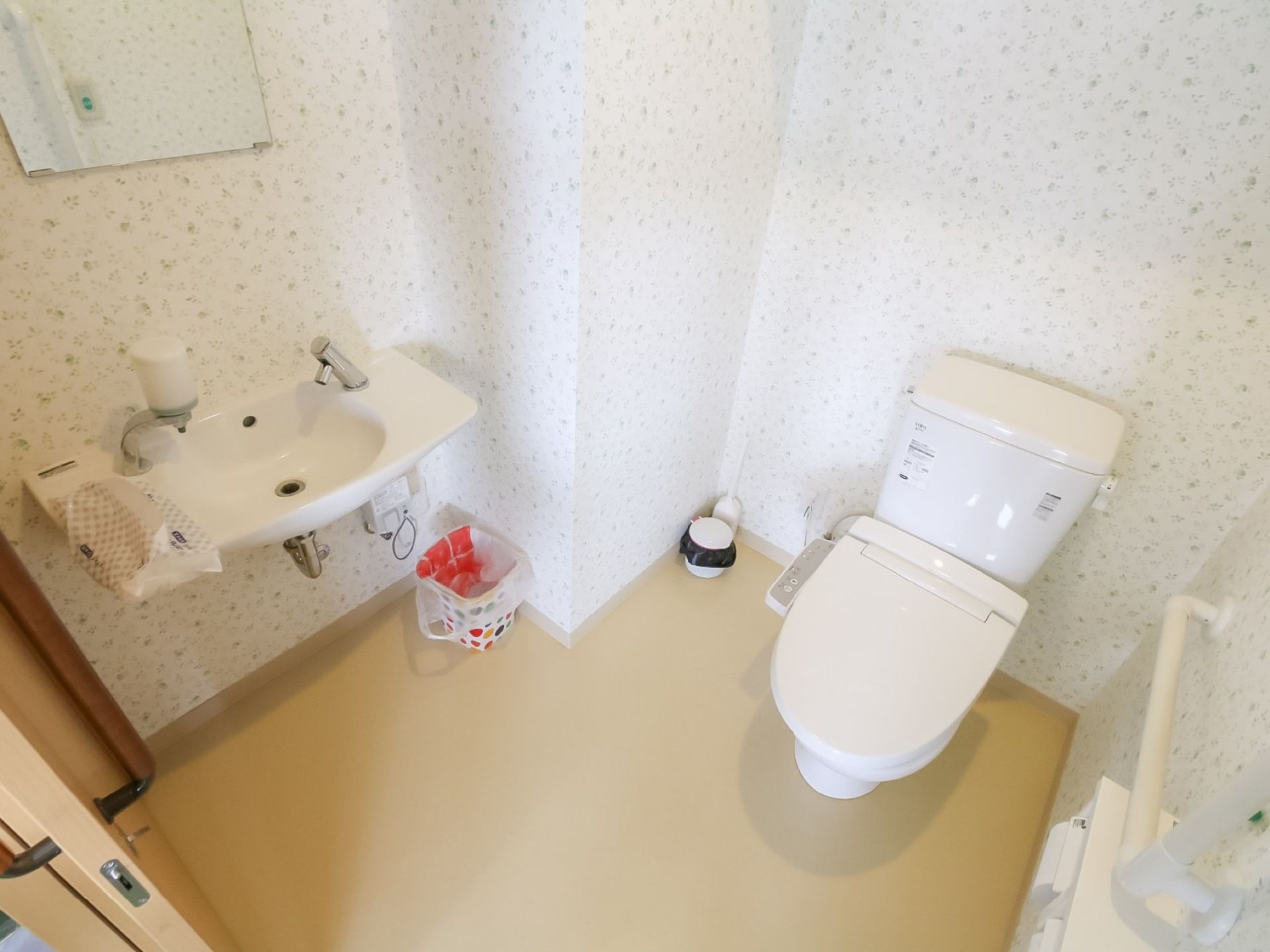 細かい模様の壁紙の広い個室のトイレがある。便器には蓋が付いている。側には鏡と洗面台があり、壁には手すりが設置されている。
