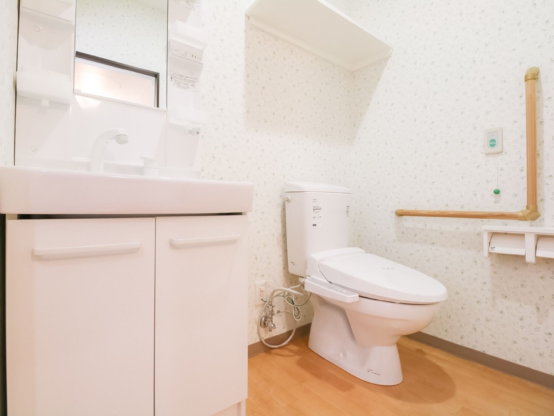 フローリングの広いトイレがある。側には棚と鏡の付いた洗面台がある。便器には蓋が付いており、側には手すりが設置されている。