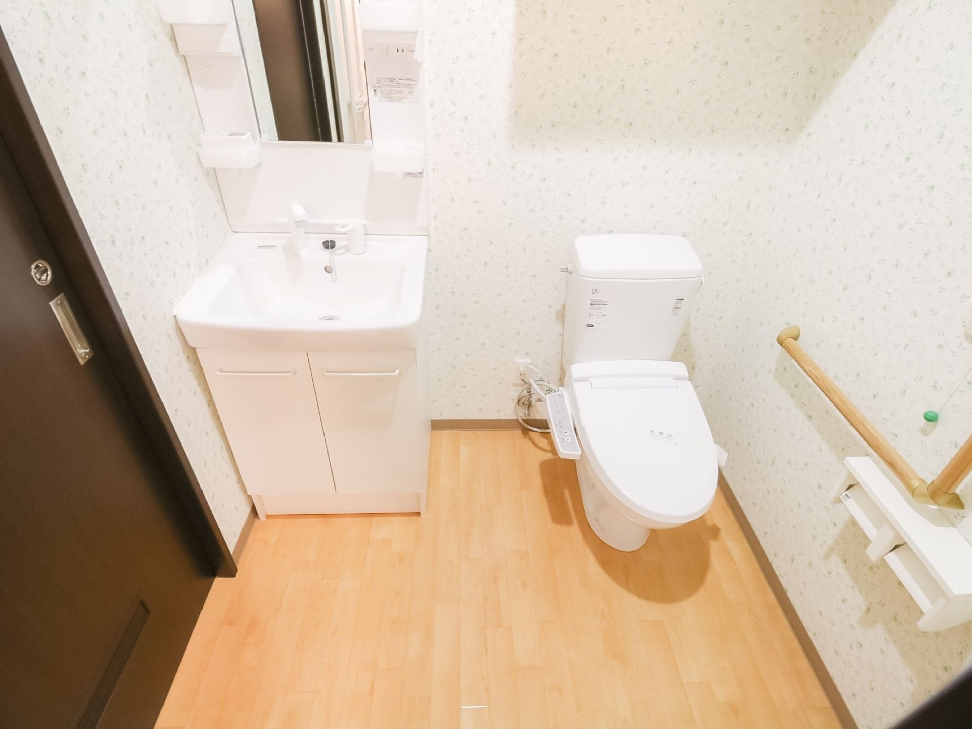 白い壁とフローリングの広いトイレと洗面台がある。洗面台には大きな鏡と小物が置ける棚があり、下部分には収納棚が付いている。