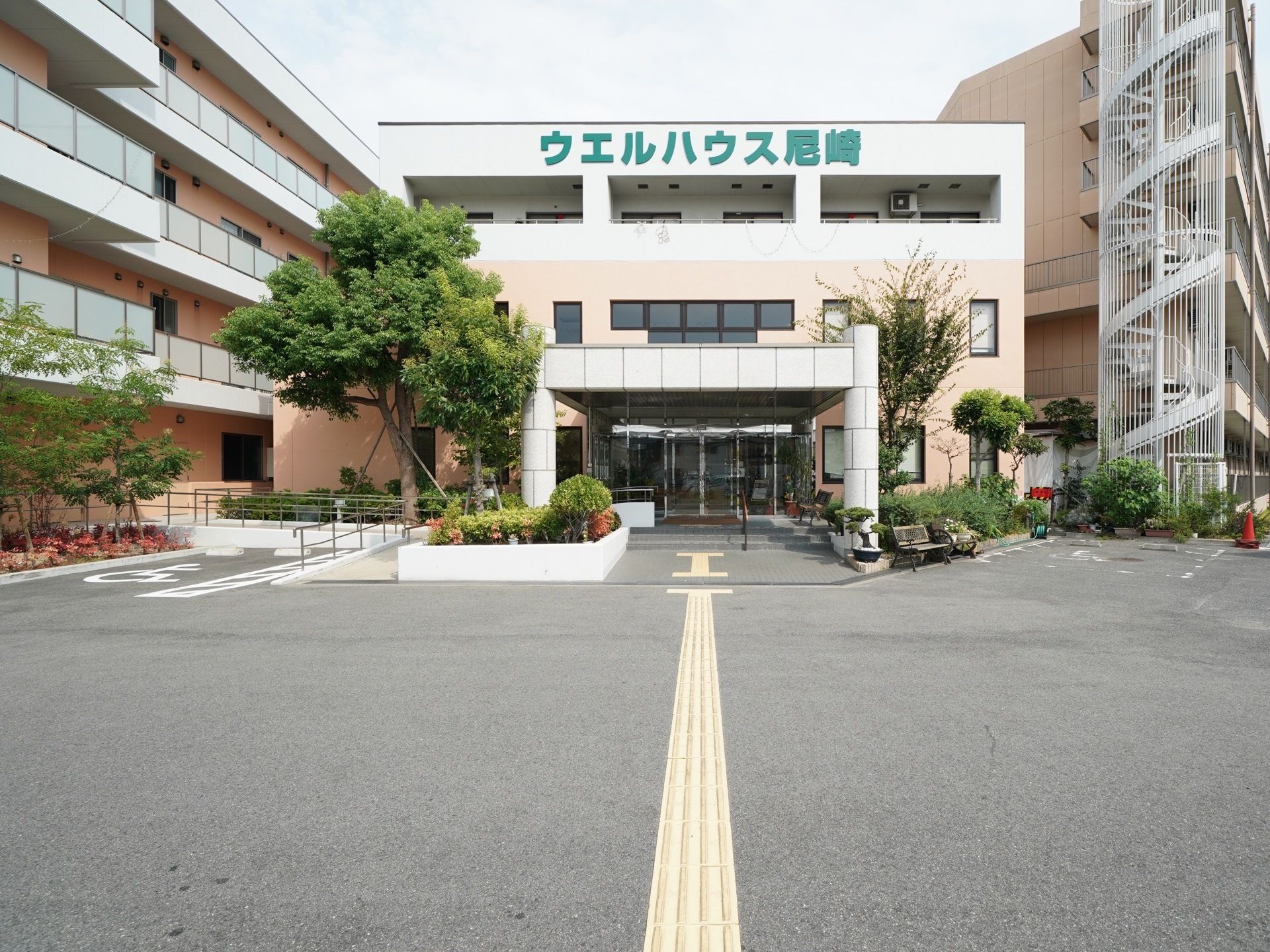 正面玄関の左右に居室エリア広がる。玄関には車椅子も通れるスロープもあり、広い自動ドアで出迎える。また、駐車場スペースも完備している。