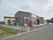 周りにあまり住宅のない閑静な土地に、濃い茶色い壁の平屋建てが建っている。施設名の看板が赤い文字で掲げられ、建物の前には自動販売機がある。