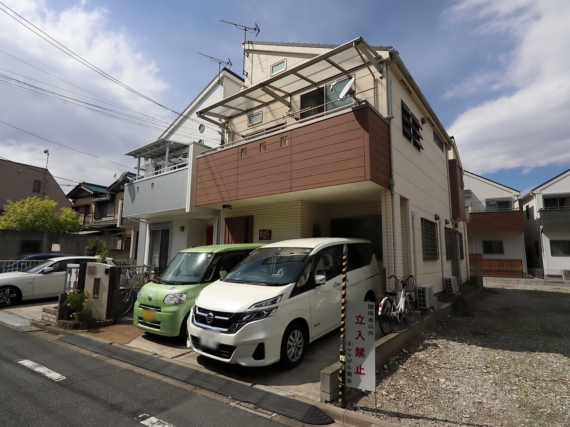 施設の外には車が停められており、２階にはひさし付のバルコニーがある。ガレージと道路の間には段差を埋めるブロックが設置されている。